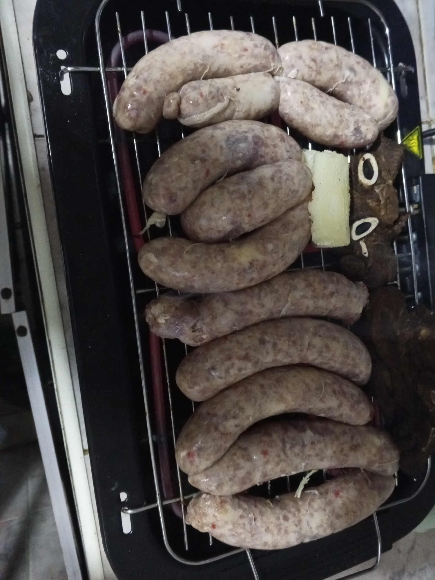 The image shows an electric grill with several sausages of varying shades of gray and a few pieces of dark-colored meat. Next to the meat appears to be a small piece of white cheese. The photograph was likely taken in Tinaquillo, Venezuela.