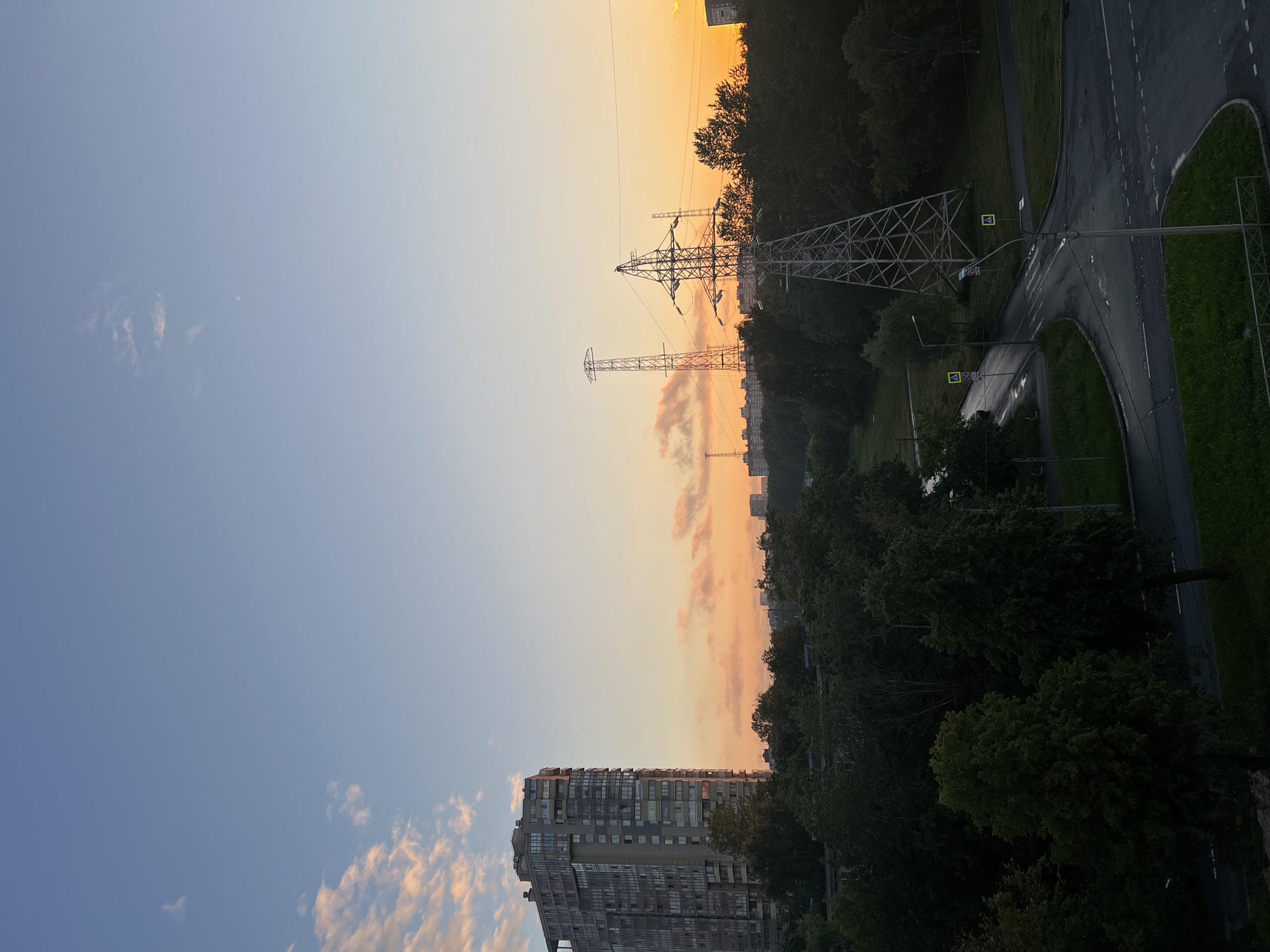 This is an image of a residential district in Saint Petersburg, Russia, taken at sunset. The sky is a gradient of blue and orange, with some wispy pink clouds. In the foreground, on the left side of the image, is a tall, modern residential building with many windows. In the middle ground, there is a dense cluster of green trees. To the right of the trees, there are two large metal electricity pylons. A road intersection with streetlights and yellow pedestrian crossing signs is visible in the lower right portion of the image, appearing wet, suggesting recent rain. In the background, more buildings and trees are visible, fading into the distance under the colorful sunset sky. The overall scene depicts a typical urban landscape at dusk.