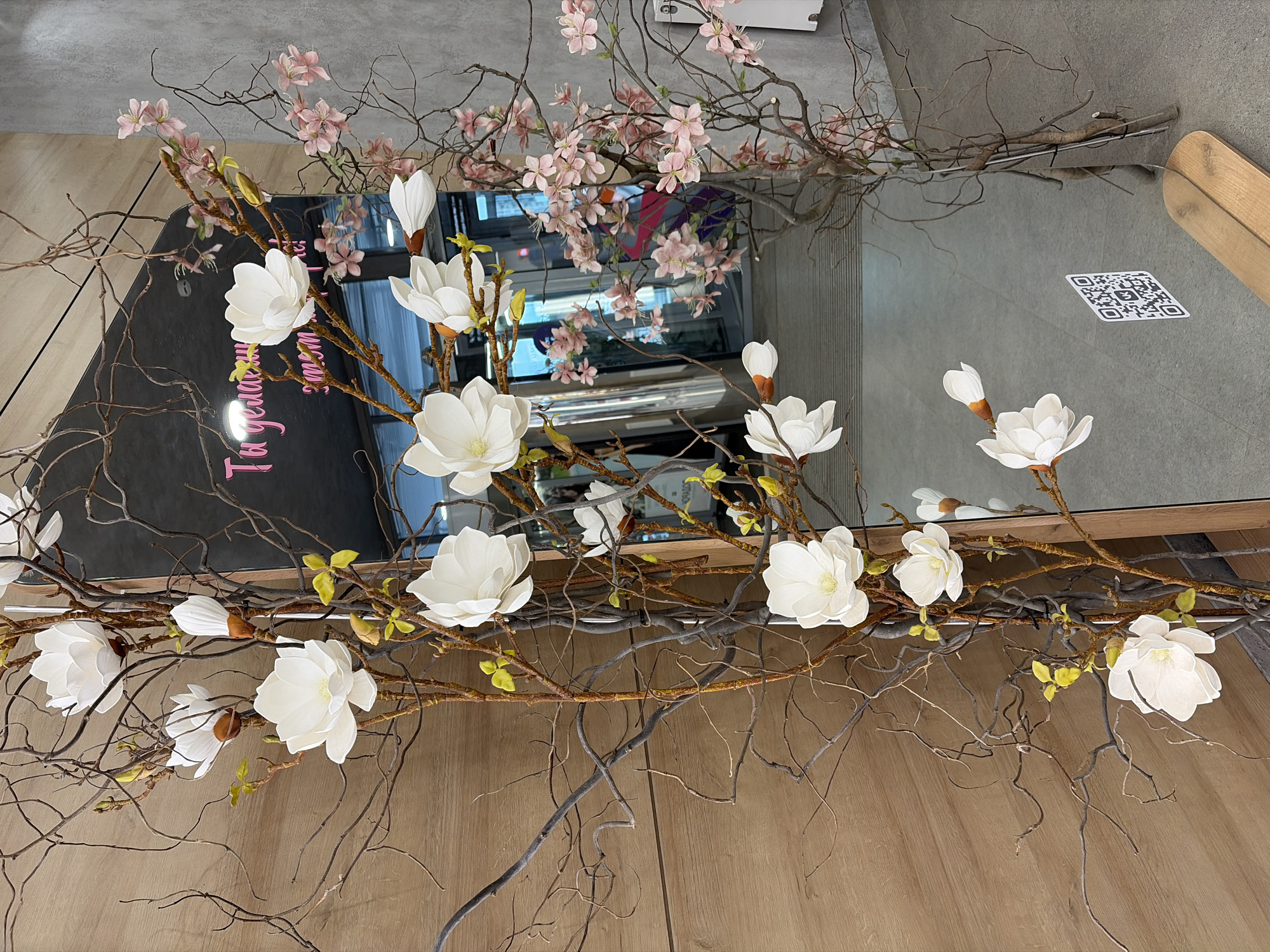 The image displays an indoor decorative arrangement featuring artificial botanical elements and a large mirror, situated within a commercial or public space in Izhevsk, Russia.
To the left of the frame, a textured light brown wooden paneled wall serves as a backdrop. In the foreground, numerous dark brown, gnarled bare branches are adorned with multiple large, white artificial flowers resembling magnolias. Interspersed among these branches are small clusters of bright yellow-green leaves or developing buds.
A large, vertically oriented mirror occupies the central and right portions of the image. Its surface reflects elements of the surrounding environment. Prominently reflected in the upper-central part of the mirror is a pink, cursive Cyrillic inscription that reads "Ты делаешь этот мир лучше," translating to "You make this world better." Blurred reflections of at least two individuals are discernible within the mirror, one in the upper-central region and another further down, partially obscured by floral elements. A white square sticker with a black QR code, featuring the VK (Vkontakte) logo, is affixed to the lower-left area of the mirror.
To the right of the mirror, a light grey concrete or textured plaster wall is visible. In the foreground on this side, similar bare branches are decorated with numerous light pink artificial flowers, resembling cherry blossoms.
The floor, visible directly and in reflection, is a light grey tiled or concrete surface. At the very bottom right, a segment of a light-toned wooden plank or bench is partially visible, extending beneath the mirror. The overall scene suggests a contemporary interior design intended for aesthetic appeal.