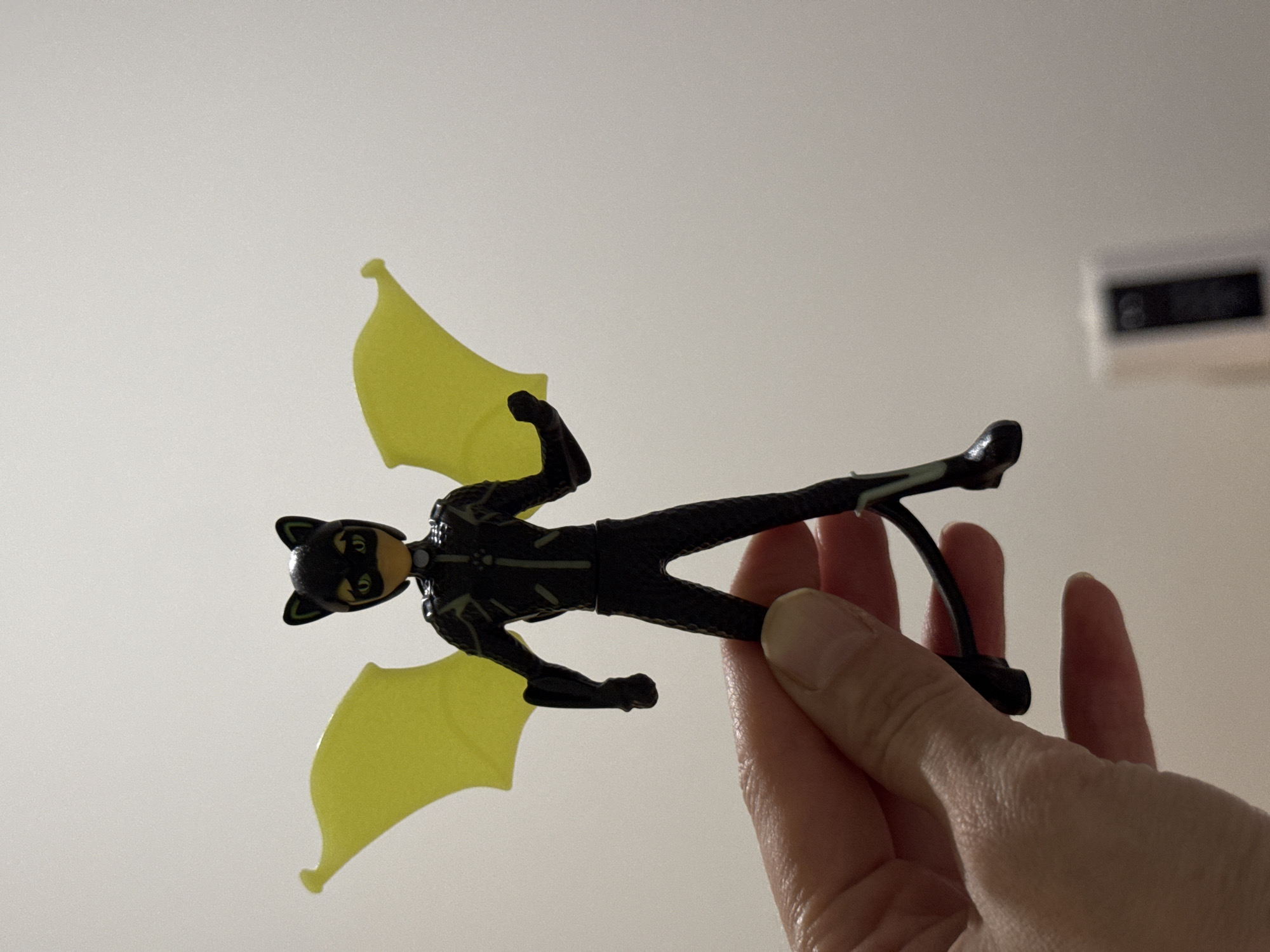 An upward-facing shot shows a human hand holding a toy figure against a plain, light-colored background, likely a wall or ceiling indoors. The toy figure depicts a character resembling Cat Noir from Miraculous Ladybug, dressed in a black full-body suit with subtle light green or yellow outlines and markings. The figure wears a black mask with prominent cat ears and has two large, translucent yellow wings attached to its back, suggesting it is in a flying pose. The hand holds the figure by its lower leg and foot, positioning it vertically in the frame.
In the upper right background, a blurred, dark rectangular object is partially visible, possibly an air vent or part of an electronic device on the wall. The lighting is even and soft, typical of an indoor setting. There is no visible text in the image. The scene takes place indoors in Izhevsk, Russia.