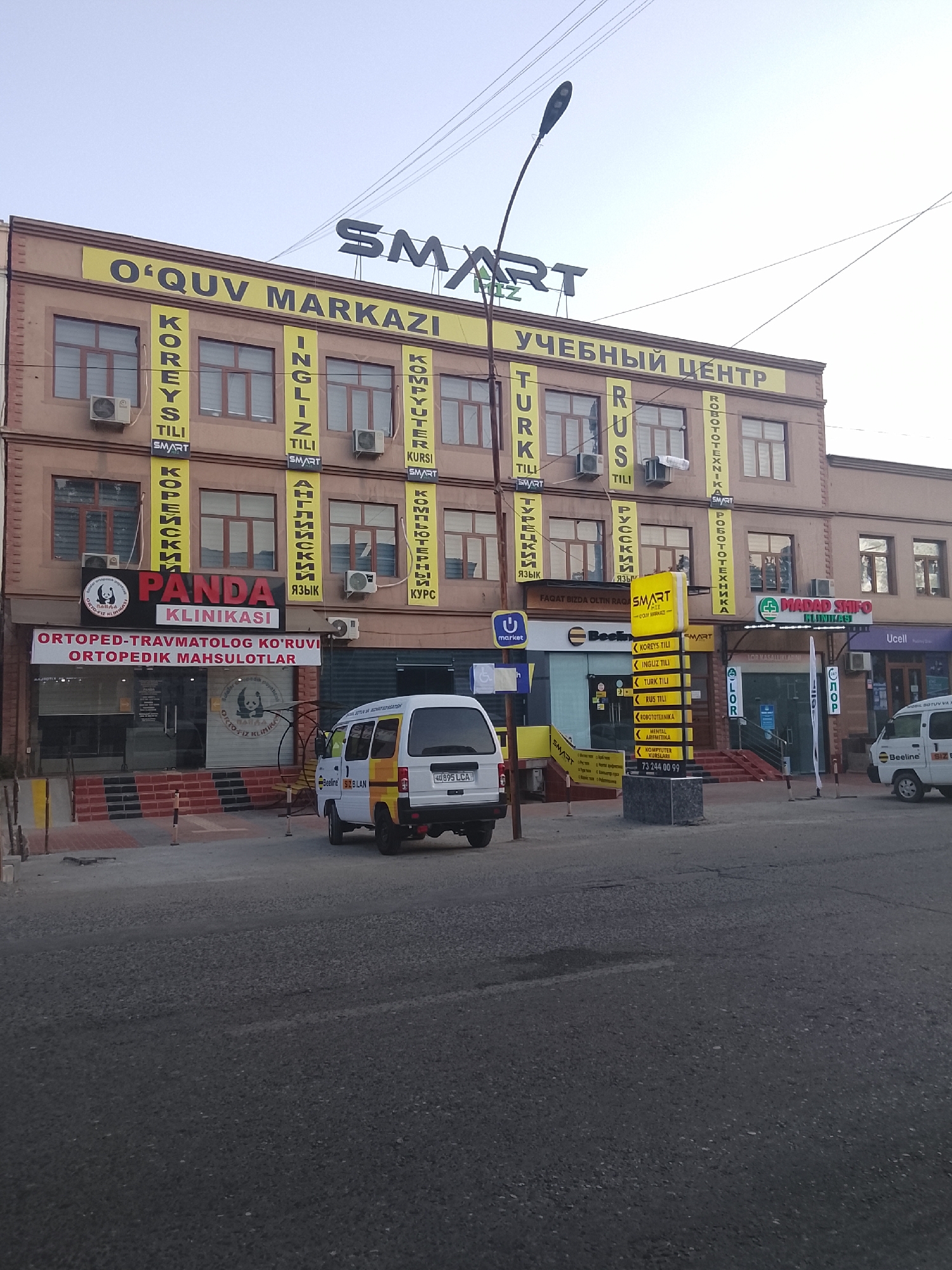 The image shows the exterior of a multi-story building in Fergana City, Uzbekistan, likely a commercial or educational establishment. The building features a light brown facade with large windows and air conditioning units visible on several floors. Prominent signage in yellow and black banners displays various languages, including Uzbek, Russian, and English, indicating it's an educational center. Some signs read "O'QUV MARKAZI" (Education Center), "УЧЕБНЫЙ ЦЕНТР" (Educational Center), and lists of languages such as "KOREYS TILI" (Korean Language), "INGILIZ TILI" (English Language), "TURK TILI" (Turkish Language), and "RUS TILI" (Russian Language).
On the ground floor, a clinic or medical center is visible with a sign reading "PANDA KLINIKASI" (Panda Clinic) and "ORTOPED-TRAVMATOLOG KO'RUVI" (Orthopedic-Traumatology Examination). Next to it, there are storefronts with entrances. In front of the building, a white van with yellow and blue accents is parked, bearing the license plate "H0895LCA". Another white van is partially visible further down the street. The street in front of the building is paved with asphalt, and there are sidewalks and streetlights. The overall impression is that of a busy urban street scene in Fergana City.