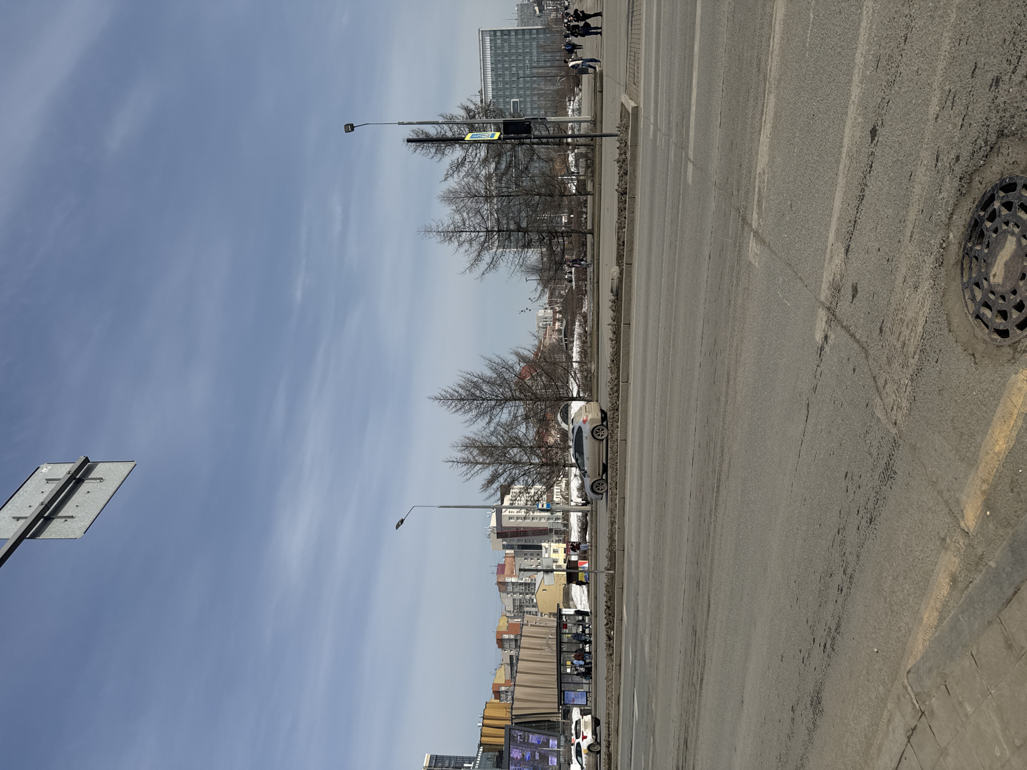 This image depicts an outdoor, daylight urban street scene on Lenin Street in Perm, Russia, likely near the Tsentralnyy Rynok (Central Market) or Komsomolskaya Square area.
The scene is dominated by a wide, multi-lane asphalt road in the foreground and midground, appearing dry but soiled with dirt and tire marks. A black, circular manhole cover is visible in the lower right corner, next to a section of concrete sidewalk with a yellow-painted curb line.
On the left side of the street, a white Yandex.Taxi vehicle is stopped near a modern bus shelter with a dark frame and glass panels, where several individuals are waiting or seated. Behind the bus stop, a multi-story building with a distinctive, multi-colored (orange, brown, grey) wavy or corrugated facade is prominent, adjacent to a modern Sberbank building featuring large glass panels and a digital display.
Across the road, a beige SUV, appearing dusty, is either parked or moving slowly. A median strip with bare deciduous trees (approximately five, possibly larch) and visible dirt/debris separates road sections. A tall, dark street light pole stands on the right, bearing a yellow pedestrian crossing sign.
In the background, a large, modern glass-fronted office building is visible on the far right. A cluster of older residential buildings with colorful facades (pinks, yellows, blues) is discernible behind the commercial complex on the left. Numerous pedestrians are present on sidewalks on both sides of the street, dressed in typical urban clothing, engaged in walking or waiting.
The sky is a clear light blue with scattered wispy clouds. A grey, rectangular road sign is partially visible in the upper left corner, attached to an overhead gantry. The bare trees and ground conditions suggest a late winter or early spring environment.