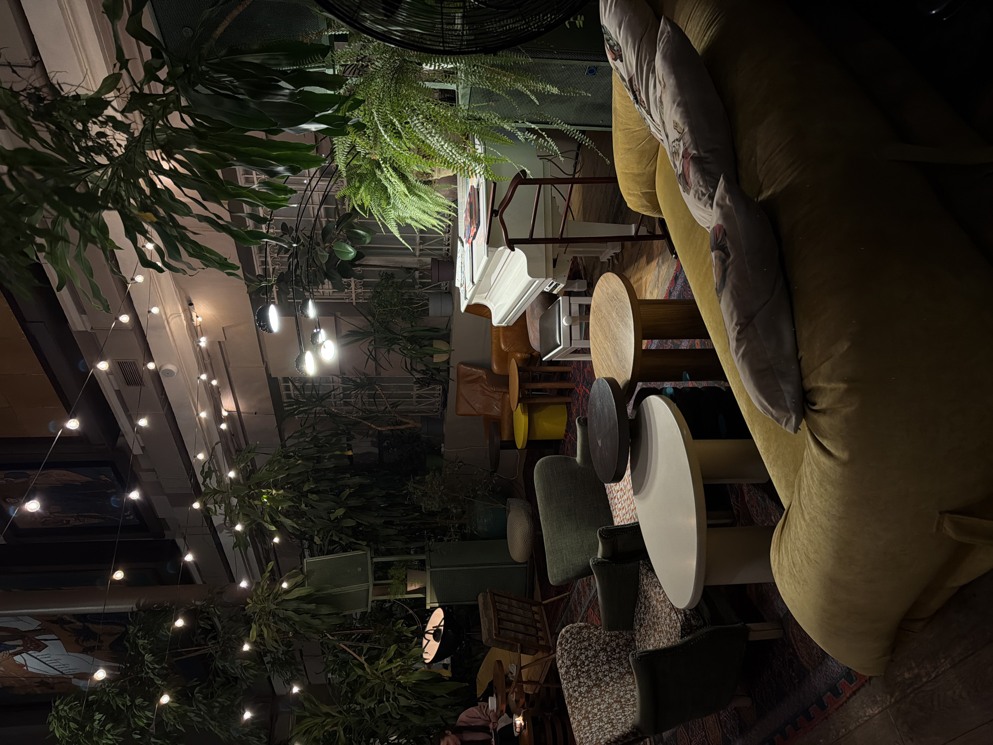 The image depicts an indoor scene, likely a lounge or cafe, with a bohemian and eclectic atmosphere. Dominating the frame is a large, plush, mustard-yellow sofa adorned with decorative pillows. In front of the sofa, several round tables of varying heights and materials are arranged, accompanied by a mix of seating options including green upholstered chairs, patterned chairs, and wooden stools. A white upright piano stands to the left, its lid open, suggesting it might be used for entertainment. Lush greenery, including ferns and large-leafed plants, is scattered throughout the space, hanging from above and placed in pots, creating a natural and vibrant feel. String lights with exposed bulbs are suspended from the ceiling, casting a warm, ambient glow, indicating it is likely evening or nighttime. The scene is well-lit, with a focus on the cozy and inviting nature of the space. There is no visible text in the image. The overall impression is one of a relaxed and artistic environment, possibly a popular spot for socializing or enjoying a quiet evening.