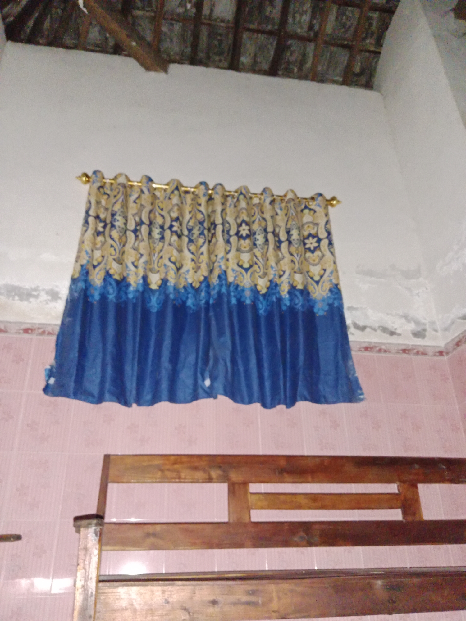 The image shows an interior scene, possibly a bedroom or living area, in Ringinrejo, Indonesia. A blue and gold patterned curtain hangs across a window against a white wall. The ceiling above appears to be made of wood and tiles. Below the curtain, pink tiled walls are visible, and in the foreground, a portion of a wooden bench or bed frame is present. The lighting is dim, suggesting it may be nighttime or a poorly lit room. There are no people or animals visible in the image. The overall impression is of a simple, possibly rustic, domestic space.