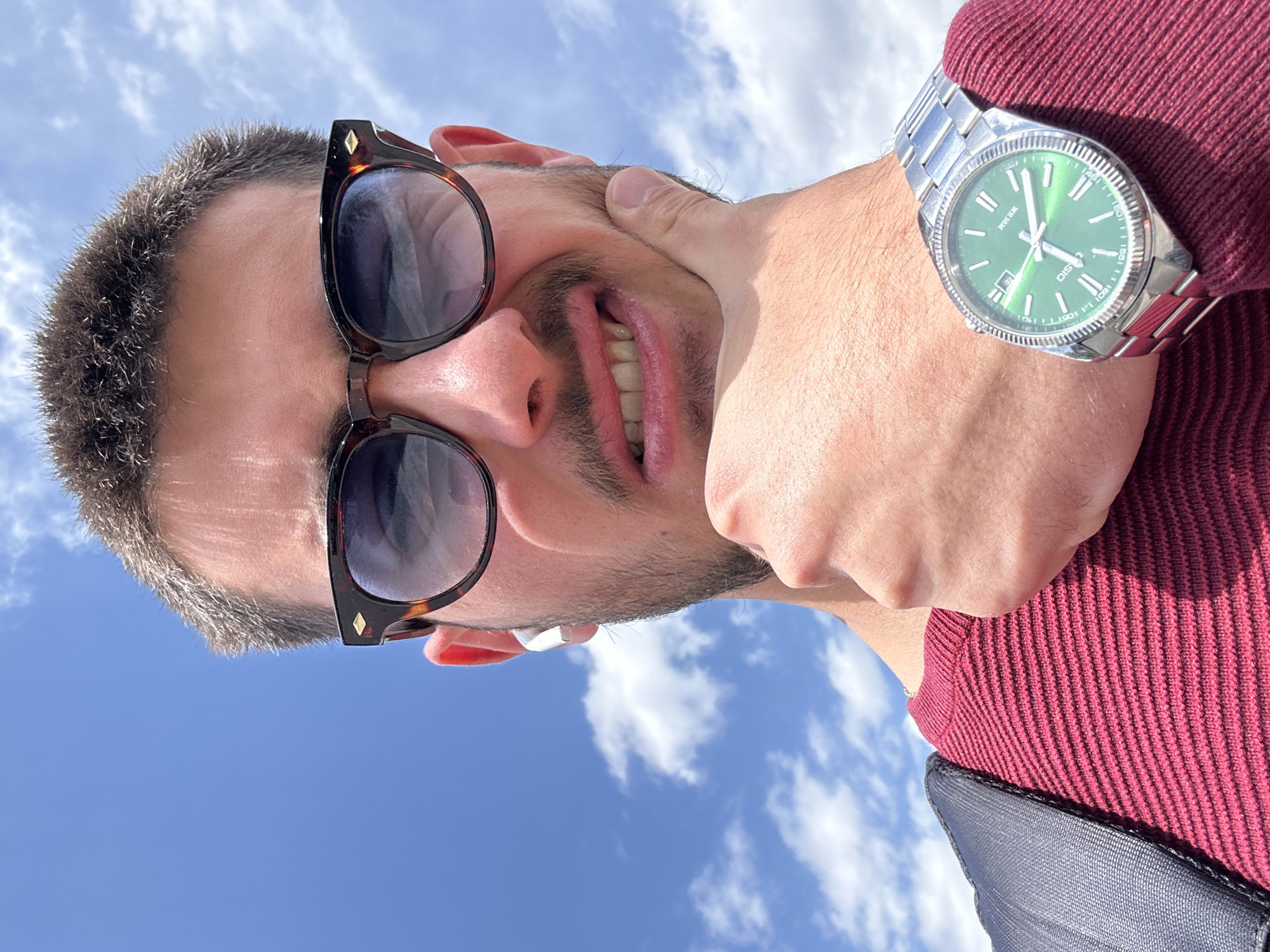 A close-up shot from a low angle depicts a man outdoors against a backdrop of blue sky with scattered white clouds. The man is wearing sunglasses with a tortoiseshell frame and a maroon sweater. A silver watch with a green face is visible on the hand that is holding the man's face. An earpiece is visible in the ear. The scene appears to be outdoors during daytime, and due to the visible Bulgarian language on the watch, the picture is likely taken in Bulgaria. It is impossible to pinpoint the exact location in Sofia without additional details.