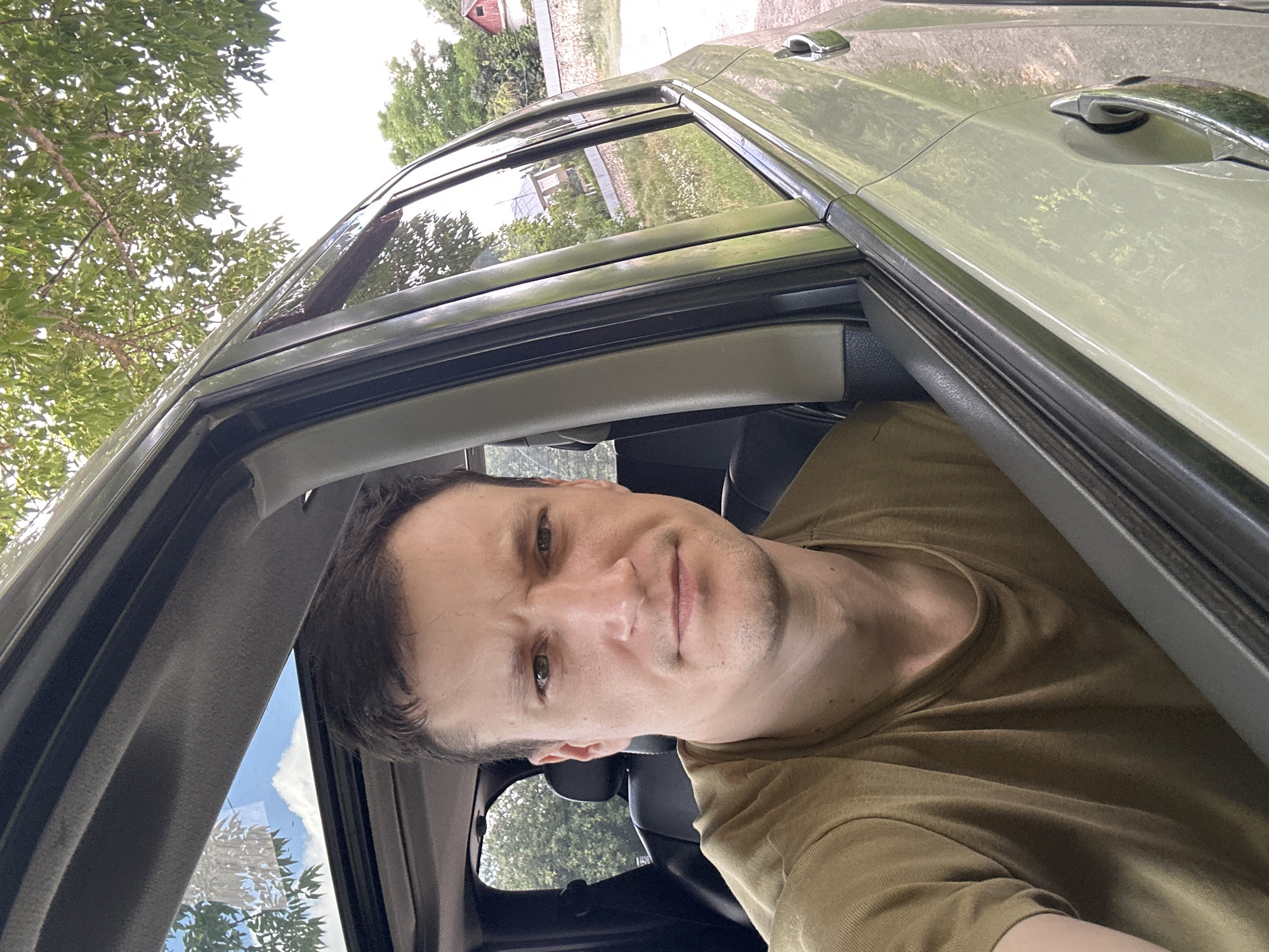 A man with short brown hair and green eyes is sitting in the driver's seat of a car, looking directly at the camera. He is wearing a short-sleeved olive green t-shirt. The car's sunroof is open, revealing a blue sky with white clouds and green tree branches. Through the car's windows, a rural landscape is visible, with trees, some buildings, and a fence. The overall scene appears to be in a village setting, possibly Makiv, Ukraine, given the provided context.