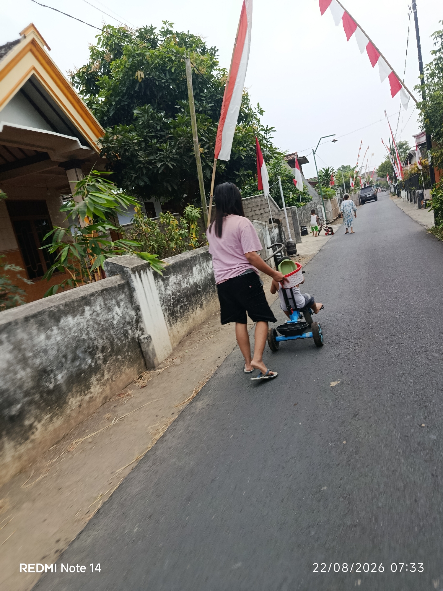 A high-angle medium shot captures a woman in a pink t-shirt and black shorts pushing a young child on a blue tricycle down a paved street. The child, wearing a gray shirt and dark shorts, is seated in the tricycle, which has a green basket on the handlebars. The woman is barefoot, wearing sandals. The street is lined with houses, some with peaked roofs and decorative trim. Red and white flags, likely Indonesian flags, are strung across the street on bamboo poles. A concrete wall, weathered and stained, runs along the left side of the street. In the background, a dark SUV is parked on the side of the road, and another person, possibly an older woman in a floral-patterned dress, walks away from the viewer. A smaller child in a white shirt is visible near the parked SUV, along with a red bicycle. Lush green trees and foliage are present along the street. The sky is overcast. The bottom left corner of the image shows the text "REDMI Note 14" and the bottom right corner displays "22/08/2026 07:33". The scene appears to be in a residential area in Panggungrejo, Indonesia, suggested by the Indonesian flags and the architecture.