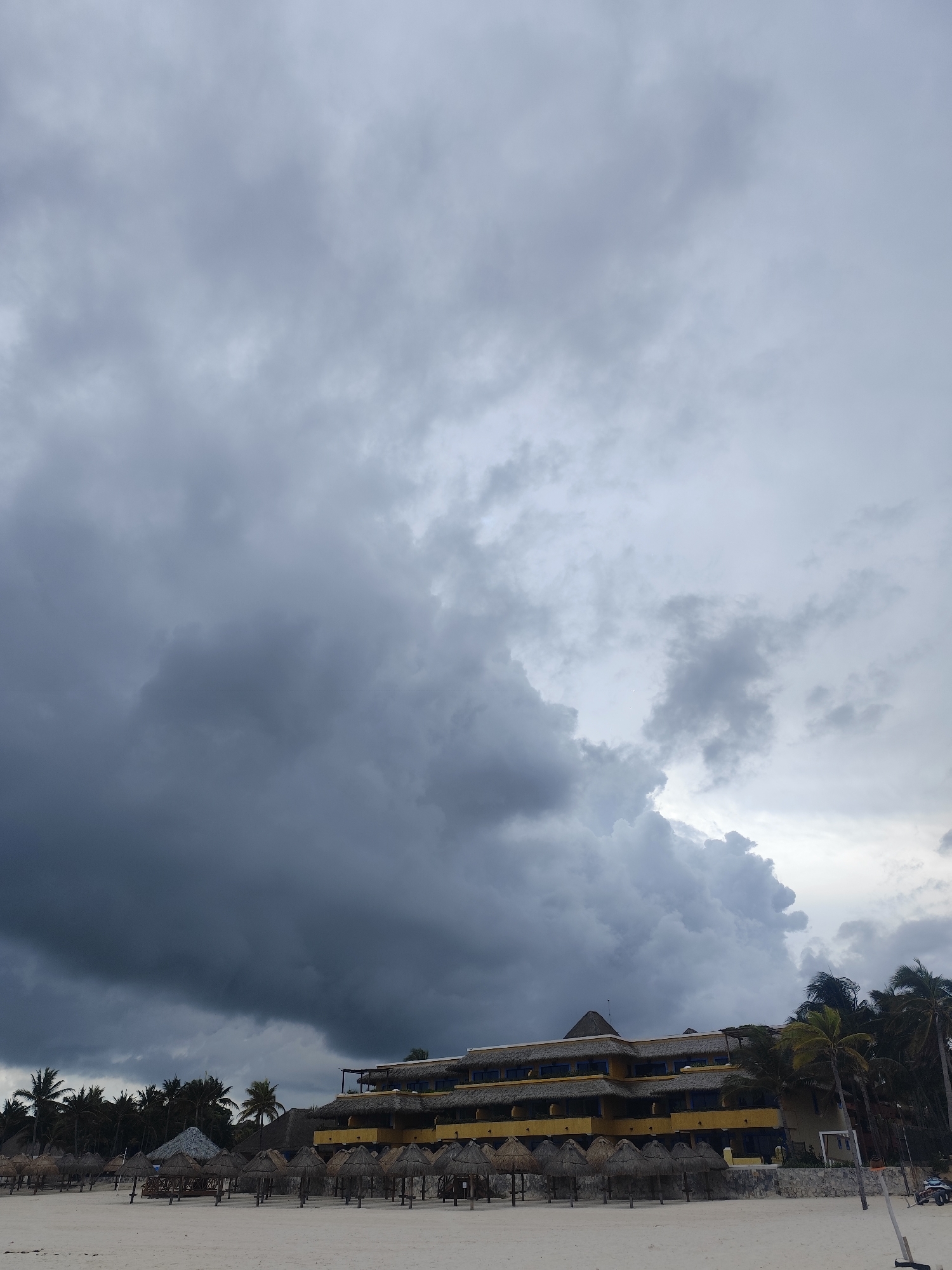 The image depicts a beach scene in Playa del Carmen, Mexico, under a heavily overcast sky. A large, dark storm cloud dominates the left side of the frame. A multi-story, yellow-painted building with a thatched roof is situated slightly right of center. In front of the building are several thatched-roof palapas lined up along the beach. Palm trees are scattered among the palapas and behind the building. The sand is light-colored. A few indistinct objects, possibly parked vehicles, are visible at the far right edge of the scene.