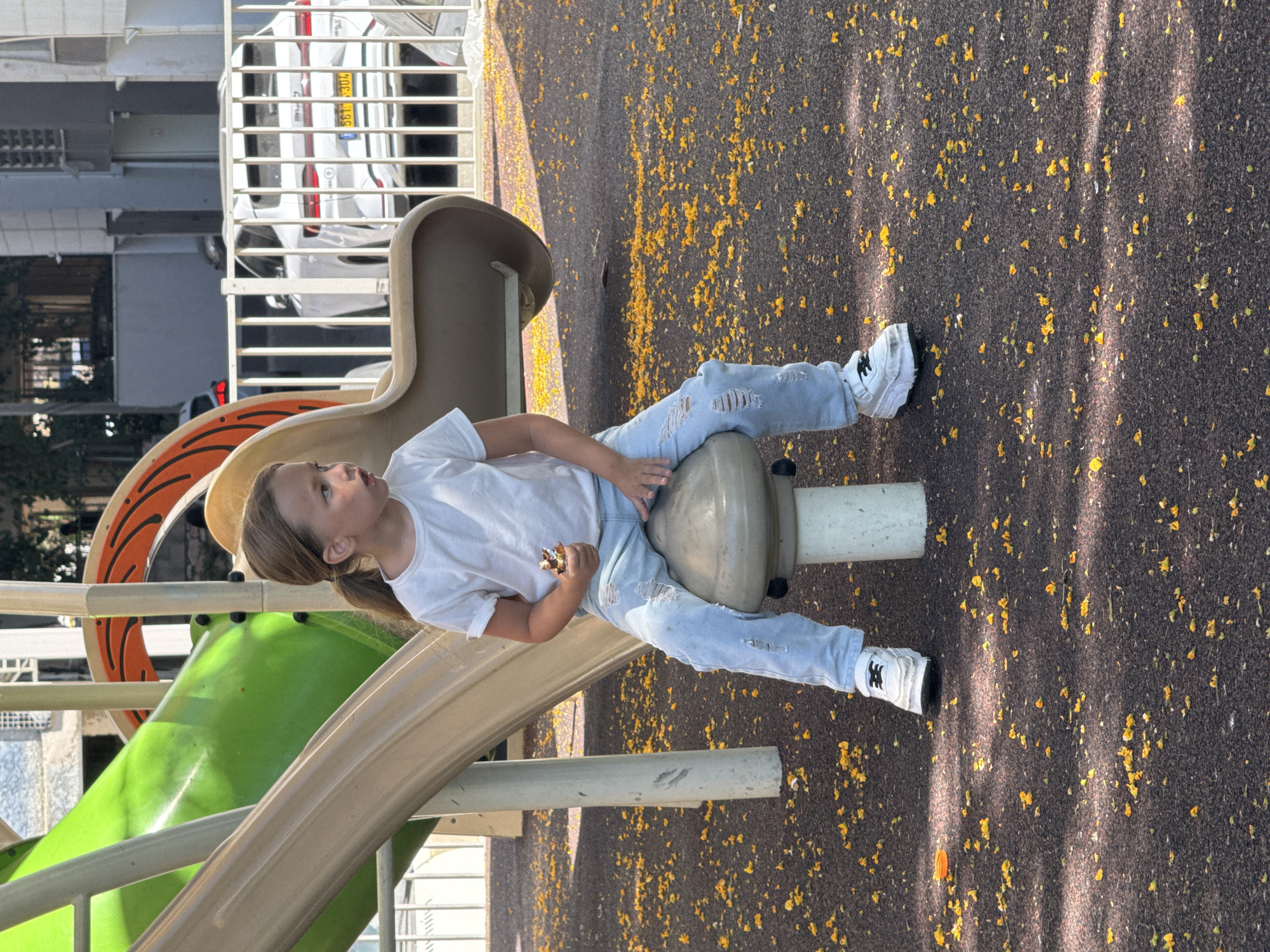A young girl with long, light brown hair tied in a ponytail sits on a mushroom-shaped play structure at a playground. She is wearing a white t-shirt and ripped light blue jeans, and white sneakers. The girl is holding a small, brown object in her right hand and looking to her right. The playground has a green and beige slide in the background. The ground is covered with dark brown material and scattered with small yellow flower petals. A white fence and cars are visible in the background, suggesting an urban setting.