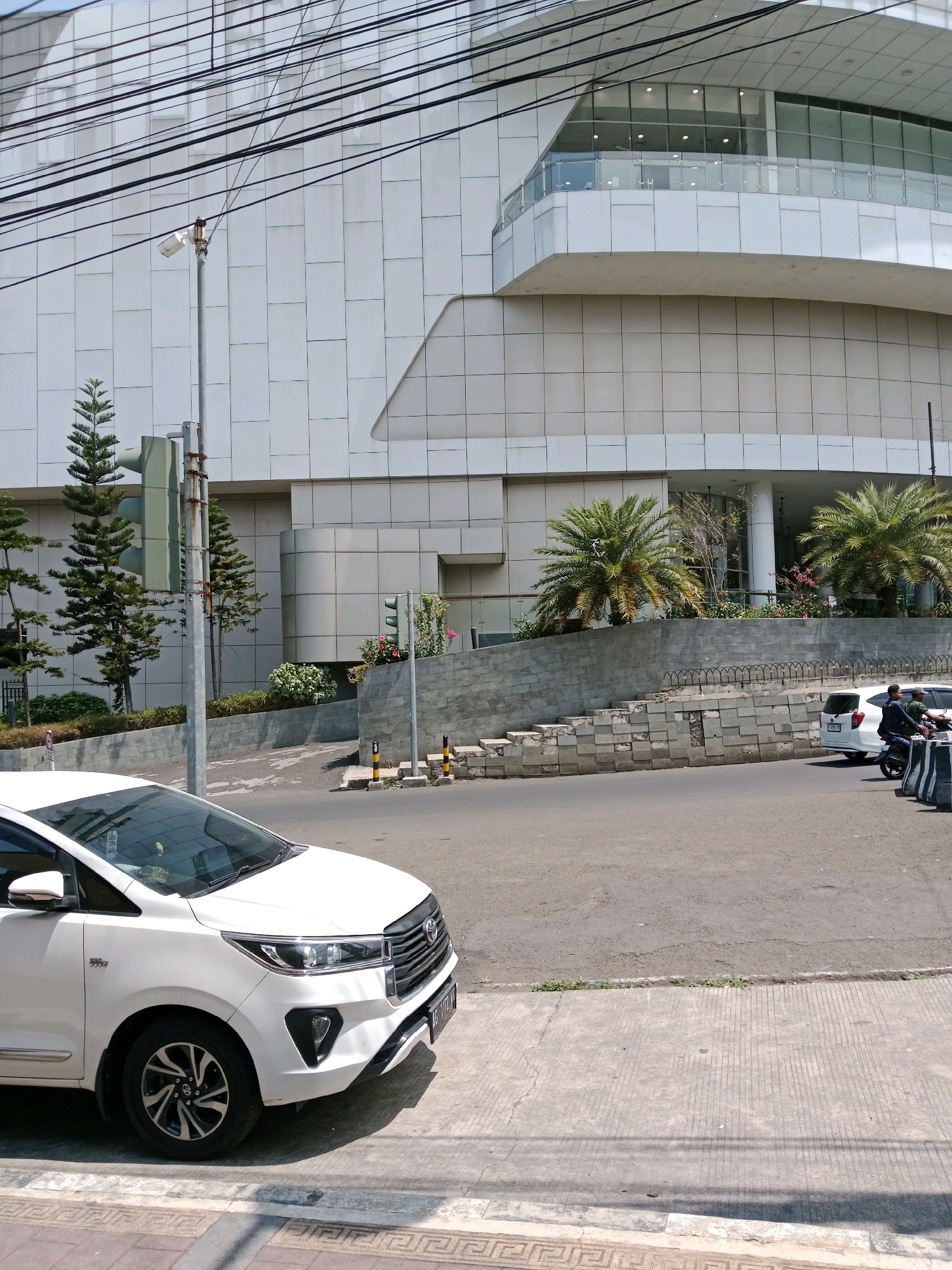 The image shows a white Toyota Innova parked on a sidewalk in front of a modern building with a white tiled facade. The building has a curved architectural feature and a balcony with a glass railing on the upper floors. Power lines are visible above the building. A street is in front of the sidewalk with a white hatchback and a motorcycle with two people on it driving away. There is a traffic light on a pole next to the building. Palm trees and other greenery are planted in front of the building. The sidewalk is made of concrete with decorative patterns. The scene appears to be in a city.