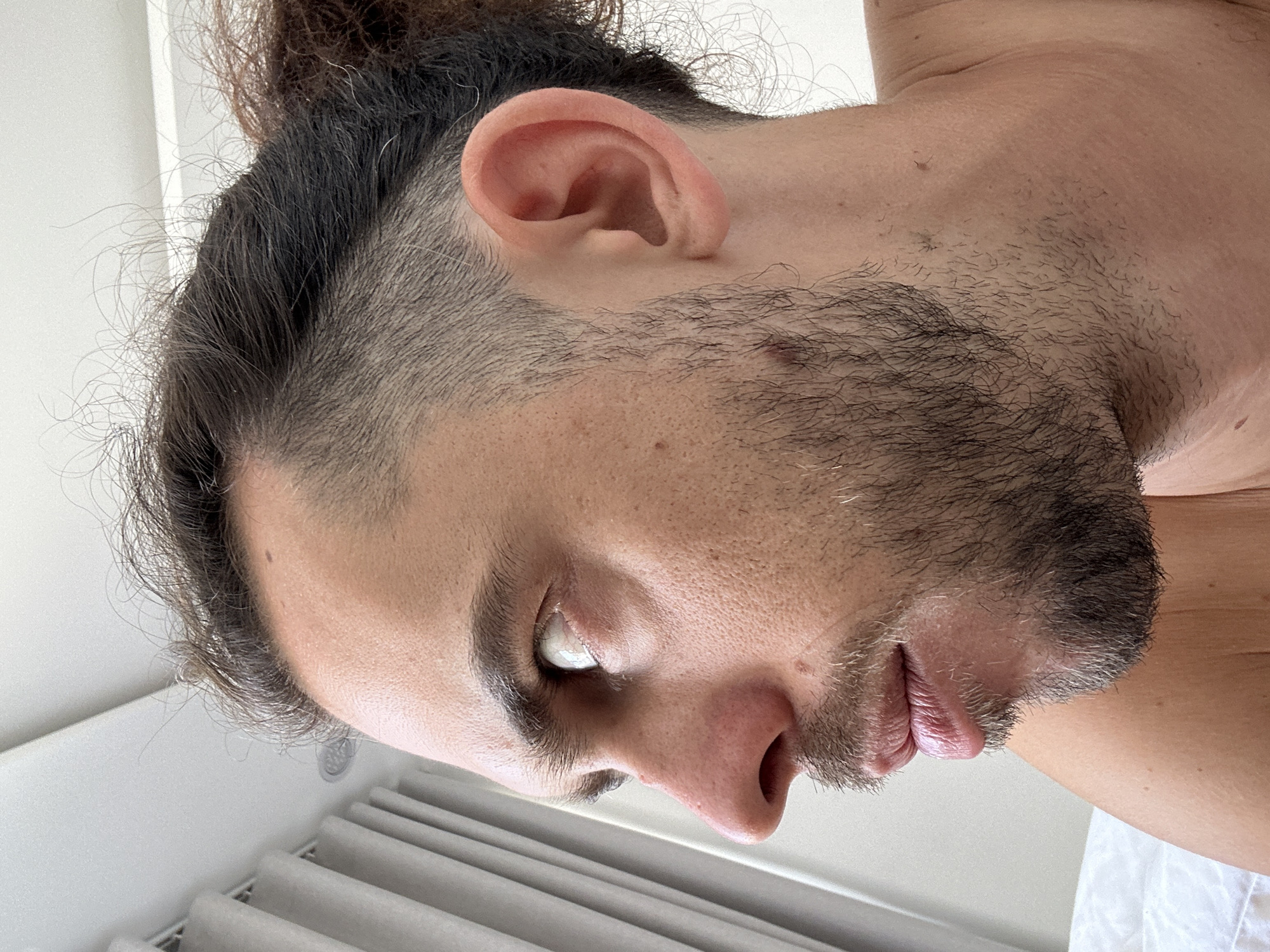 The media file is a close-up, low-angle shot of a person's face, focusing on their left side. The person has dark hair pulled back in a bun, and a light beard and mustache. Their left eye is visible, with a white sclera and a dark pupil, looking slightly upwards and to the left. The nose is prominent with visible pores. The lips are slightly parted, revealing the lower lip with a reddish-pink hue. The skin appears to have a light tan and shows signs of texture, including some light stubble on the cheek and chin. The ear is clearly visible, with a typical human ear structure. Part of the neck and shoulder are also visible, showing more light stubble and skin texture.
In the background, a white, slatted object, likely a radiator or vent, is visible at the bottom of the frame, suggesting an indoor setting. The lighting appears to be natural, possibly from a window, creating soft shadows and highlights on the face. The overall mood conveyed is neutral, with no strong emotional indicators. There is no visible text in the media file. The context provided places the scene in Đà Nẵng, Vietnam.