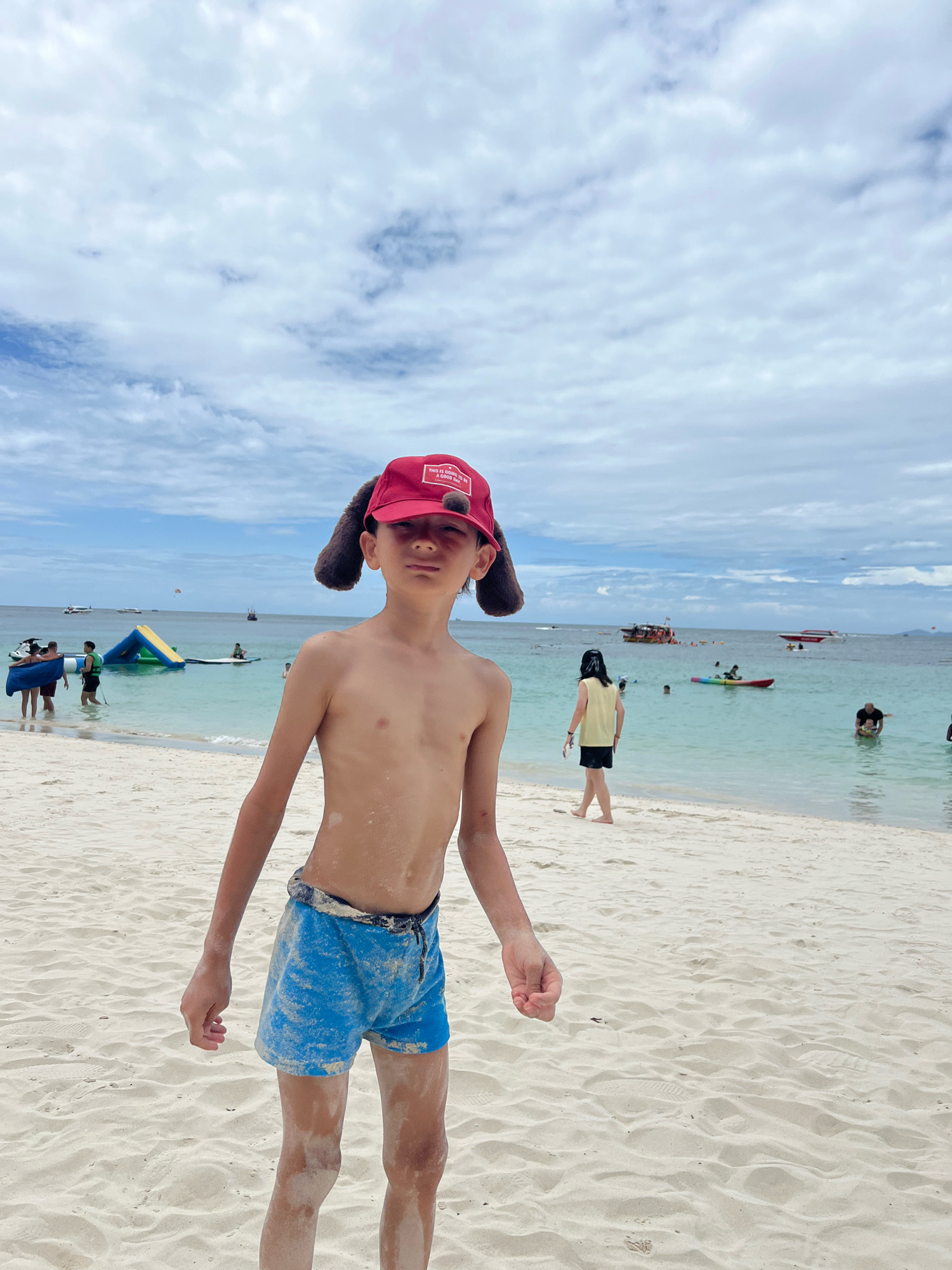 A young boy wearing a red baseball cap with dog ears and blue swim trunks stands on a sandy beach in Pattaya City, Thailand. The boy is shirtless and has sand on his body. In the background, the turquoise ocean is visible, with several boats and people enjoying water activities. The sky is partly cloudy.