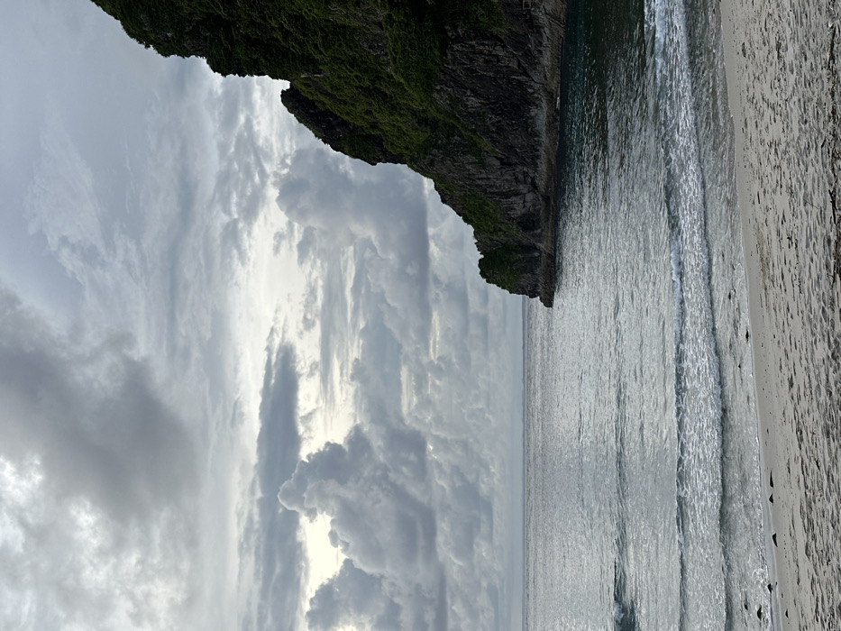 The image depicts a coastal scene in Sekongkang, Indonesia, under an overcast sky. The upper half of the frame is dominated by a dense formation of gray and white cumulonimbus clouds, with lighter patches indicating a diffused light source. The horizon line is visible across the middle, separating the cloudy sky from the expansive body of water. The water surface is a muted gray-green, showing gentle ripples and small breaking waves nearing the shore.
On the right side of the image, a steep, rocky landmass or cliff, densely covered with dark green vegetation, descends directly into the water. Its shadowed rock face is partially visible beneath the foliage. The cliff forms a curve, creating a small bay or inlet. In the foreground, a light-colored sandy beach occupies the lower left portion of the frame, extending towards the center. The sand shows faint textures and scattered dark debris near the waterline. No individuals, animals, or man-made objects are distinctly visible in the scene. The overall environment suggests a natural, undeveloped coastal landscape.