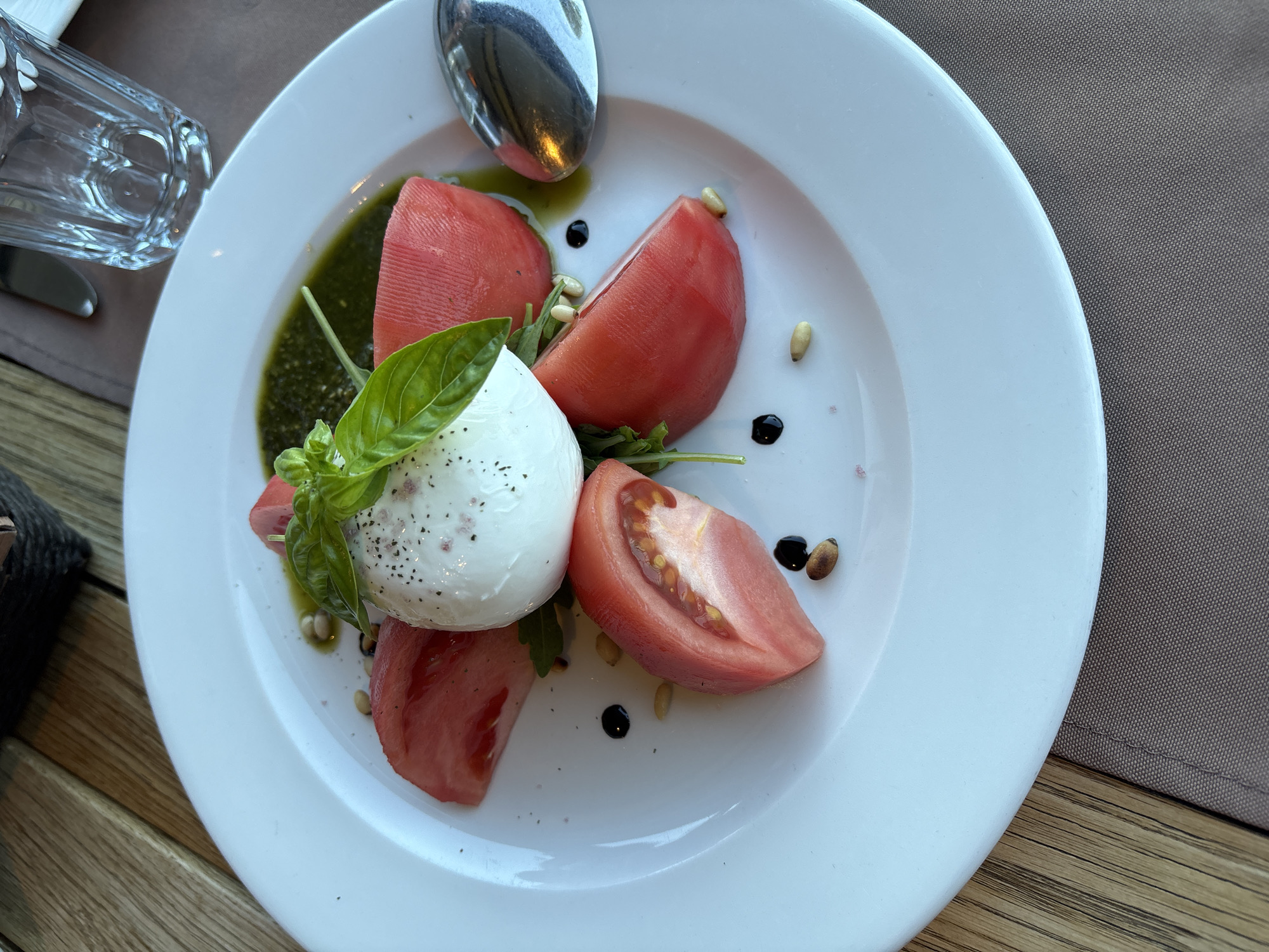 A close-up shot features a Caprese salad on a white plate, positioned on a brown tablecloth on a wooden table. The salad consists of a ball of fresh mozzarella, seasoned with black pepper, topped with basil leaves. It is surrounded by wedges of ripe tomatoes. Drizzled over the dish are balsamic glaze and a vibrant green pesto sauce, with scattered pine nuts adding texture. A silver spoon rests on the right side of the plate. In the background, a clear glass with water and part of a woven basket are visible. The setting appears to be a restaurant in Topola, Bulgaria, suggesting an outdoor or indoor dining area with natural light.