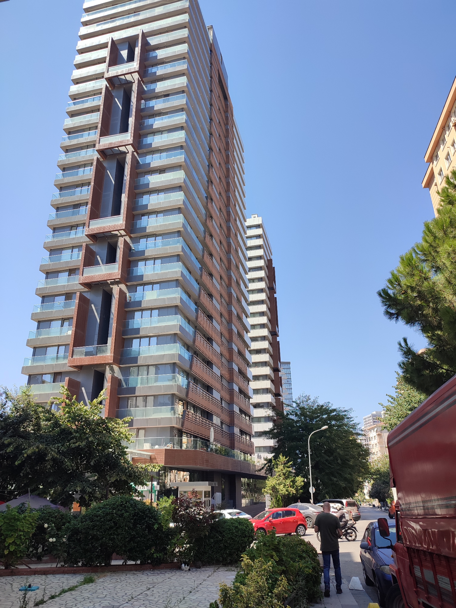A wide-angle, low-angle shot captures a street scene in Kadıköy, Turkey, dominated by a modern, tall, multi-story residential building with balconies and wood-paneled accents. The building has a textured facade with protruding sections and glass railings on its many balconies. To the right, another, slightly shorter residential building with a lighter facade is visible, partially obscured by lush green pine trees.
In the foreground, a paved walkway runs alongside a dense area of green bushes and small trees. Cars, including a bright red hatchback, a gray sedan, and a dark blue sedan, are parked or moving along the street that curves into the distance. A person in a dark green t-shirt and blue jeans stands on the sidewalk facing away from the camera, looking down the street. A motorcycle with two riders is also present on the road. A large red truck occupies the far right of the frame, with its side and rear visible. The sky is clear and bright blue. The overall impression is of a sunny day in a contemporary urban environment.