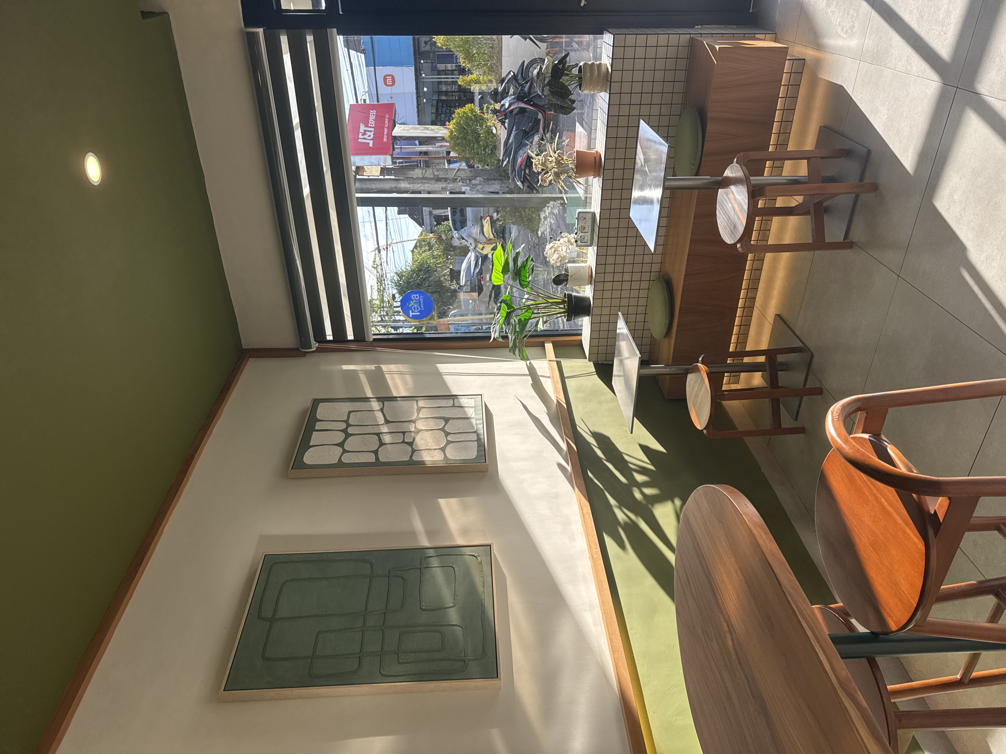 This is a wide-angle, eye-level shot of the interior of a cafe. Sunlight streams in from a large window on the right, casting strong shadows across the room. The decor is modern and minimalist, with a green ceiling, white walls, and accents of natural wood. Two framed abstract artworks hang on the left wall, both featuring geometric patterns in muted green and grey tones.
In the foreground, a round wooden table with a dark teal base is partially visible, with two wooden stools around it. To the right, a built-in wooden bench runs along the window, topped with a tiled backsplash. On the bench sit several potted plants, including a large leafy one, and a small digital clock displaying "18". Two small square tables with metal tops and square metal bases are placed in front of the bench, each with a round green cushion on the seat of a wooden stool underneath.
Through the window, a street scene is visible with a yellow scooter, motorbikes, cars, buildings, and overhead wires. Signs for "J&T Express" and "mi" are prominent. The overall atmosphere is bright and airy, with a serene, natural aesthetic.