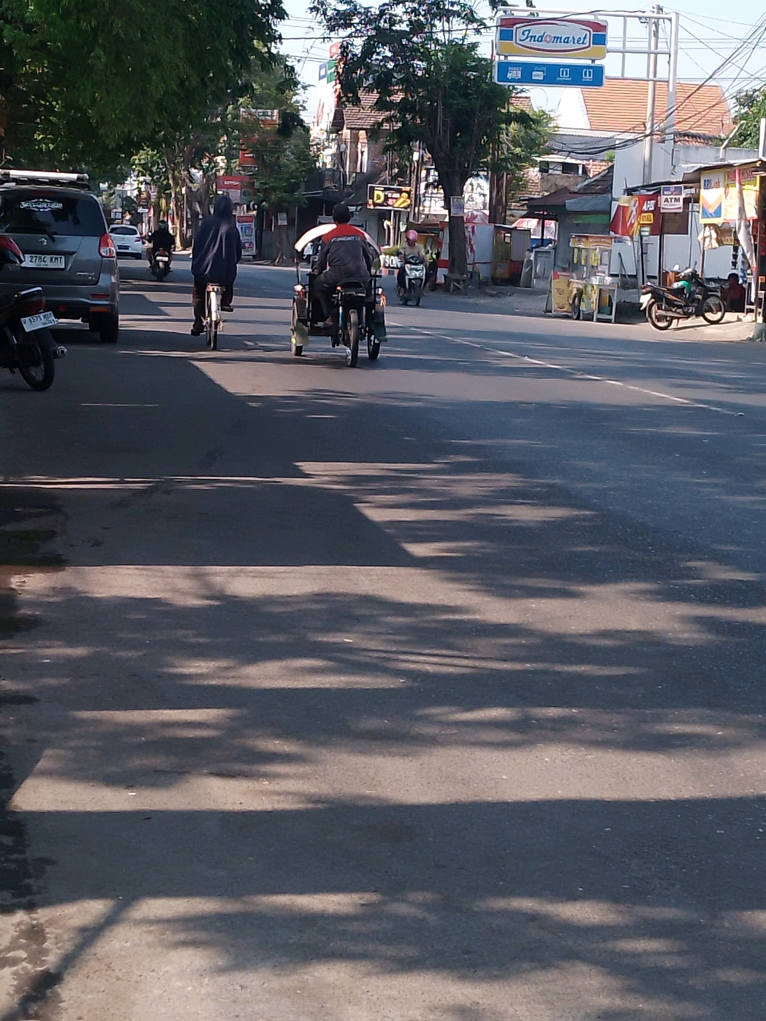 This is a photograph taken from a low angle, looking down a paved road in Kertosono, Indonesia. The scene depicts a street with moderate traffic and buildings lining the right side. In the foreground, the road surface is visible, with shadows cast by trees creating dappled patterns.
Several vehicles are present. On the left, a grey SUV is parked on the side of the road, with a portion of its rear visible. To its right, a motorcycle with the license plate "P 4577 KMI" is parked. Further down the road, a person wearing a dark blue hooded jacket is riding a bicycle. To the right of the cyclist, a three-wheeled vehicle, possibly a becak or a small motorized tricycle, is being ridden by a person wearing a grey jacket with red accents. The three-wheeled vehicle has a covered passenger area. Further down the road, another motorcycle is visible, and a few more vehicles are indistinctly seen in the distance.
The buildings on the right side of the road appear to be shops or commercial establishments. A prominent sign with the logo "Indomaret" is visible, indicating a convenience store. Other signs are also present, some displaying text that is difficult to read. Several small stalls or kiosks are set up along the sidewalk in front of the buildings. The environment suggests a sunny day, with clear skies visible between the trees. The overall impression is that of a typical street scene in a bustling town or city in Indonesia.