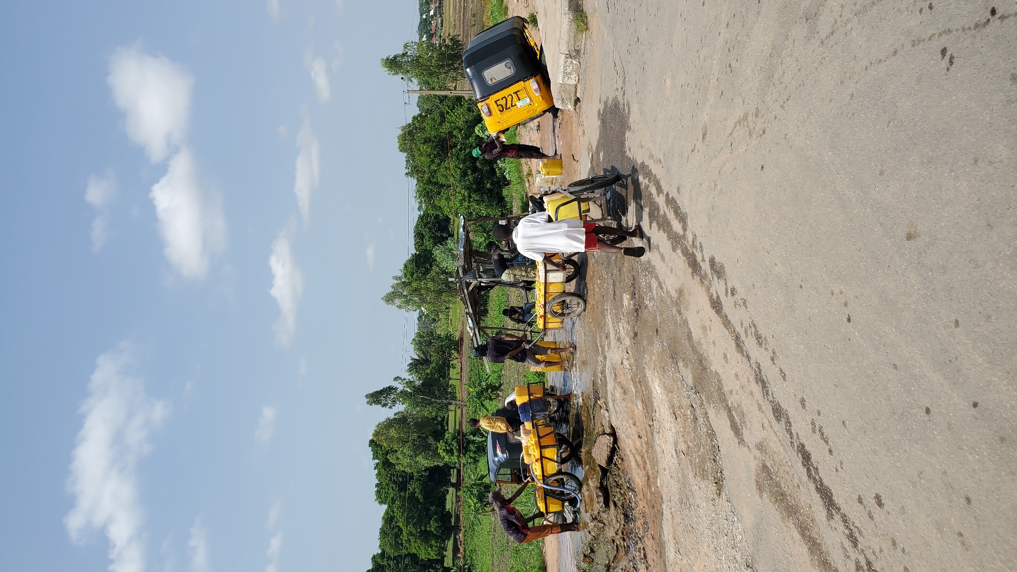 In Jalingo, Nigeria, a group of individuals is engaged in the act of filling yellow jerrycans with water. The scene unfolds on a paved road, likely a section that has been partially washed away or is prone to flooding, given the presence of water and mud. Two carts loaded with jerrycans are positioned on the left side of the road, with men attending to them, possibly using hoses to fill the containers. A third cart, also carrying jerrycans, is near a makeshift shelter on the right. Further to the right, a yellow and black auto-rickshaw is overturned, its wheels in the air, with a person in a green cap interacting with it. Another person in a white shirt and red shorts is walking away from the scene towards the foreground. The environment is characterized by lush green trees and vegetation in the background, with a clear blue sky dotted with white clouds overhead. The overall impression is one of a busy, outdoor scene where water collection is the primary activity.