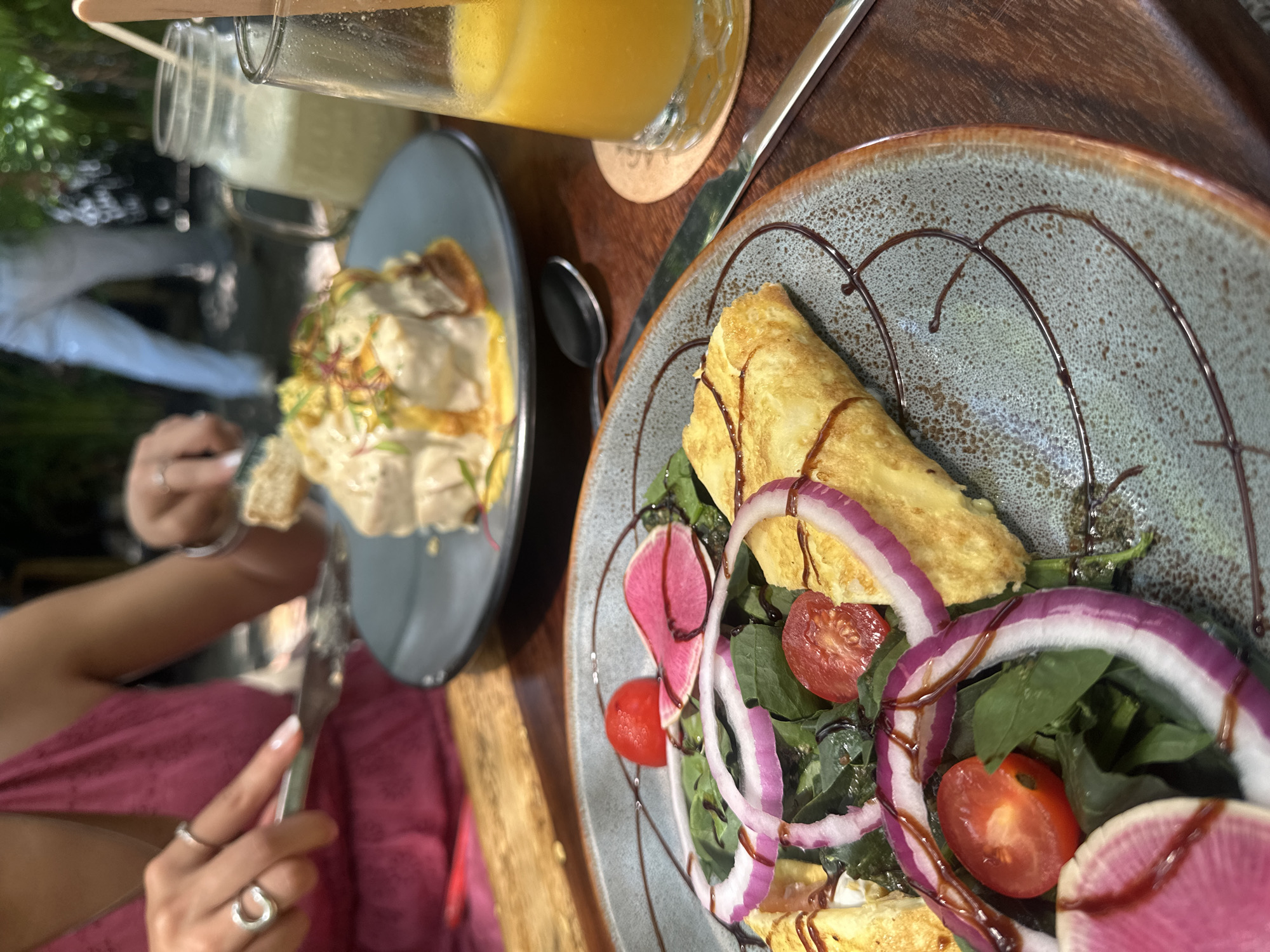A close-up shot shows two plates of food and two beverages on a wooden table. In the right foreground, a plate features what appears to be an omelet served over a bed of green leaves, with sliced red onions, tomatoes, and slices of what looks like radish on top. A dark sauce is drizzled on the omelet and plate.
To the left, there is another plate with a creamy, saucy dish topped with what looks like sprouts. A person is holding a piece of this food with their left hand and cutting it with their right.
Behind the food is a beverage in a glass jar. Next to that, in the upper right, is a glass of orange juice, with a spoon and knife.
The scene appears to be at a restaurant or outdoor cafe with vegetation in the background. It is difficult to determine if the location is in Acapulco de Juárez, Mexico, without further information or context.