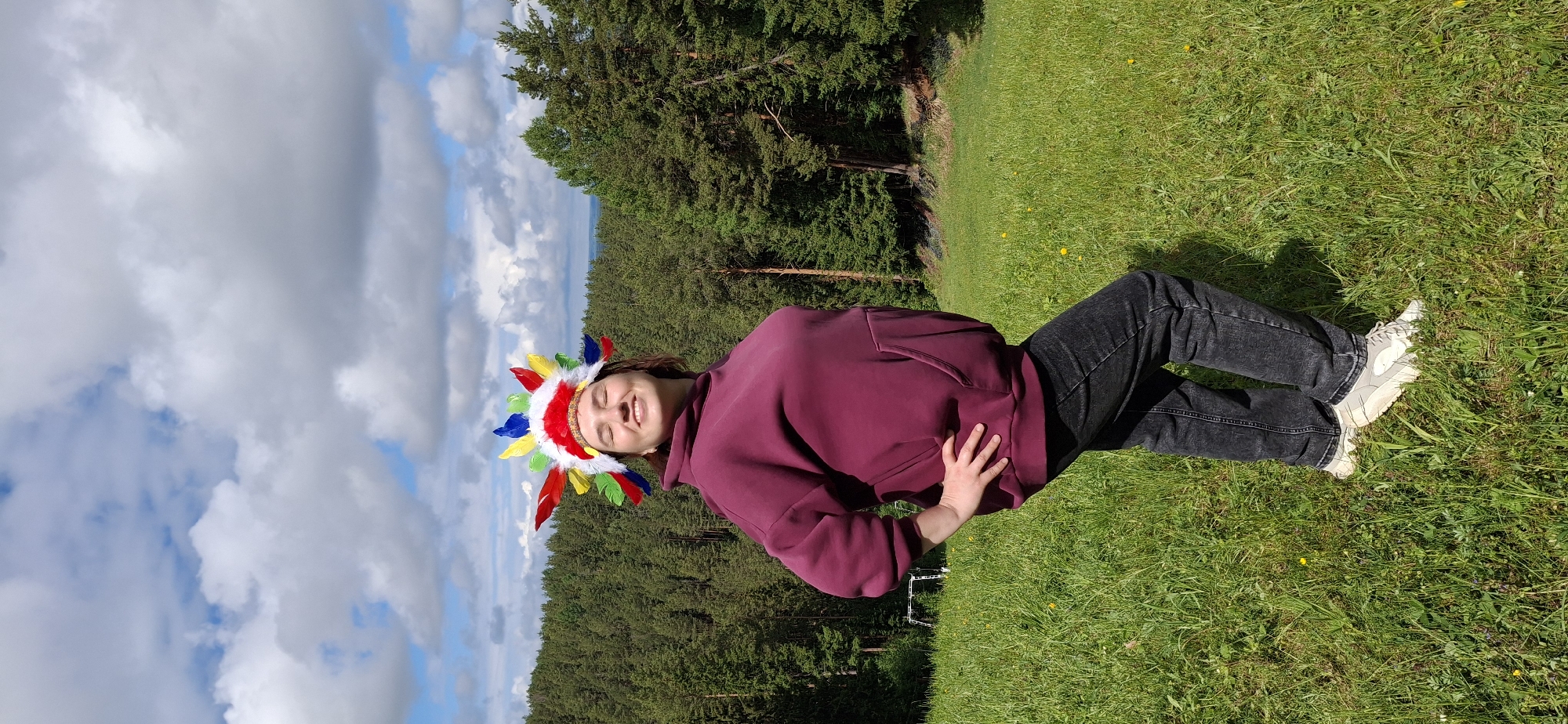 The image depicts a person lying on green grass in a park or field. The person is wearing a maroon hoodie, dark gray jeans, and white sneakers. They are also wearing a colorful Native American-style headdress with red, white, yellow, blue, and green feathers. The person's eyes are closed and they appear to be smiling or content. In the background, there is a dense forest and a cloudy sky. The location is outdoors in a natural setting. The city is Прикамский, Russia.
