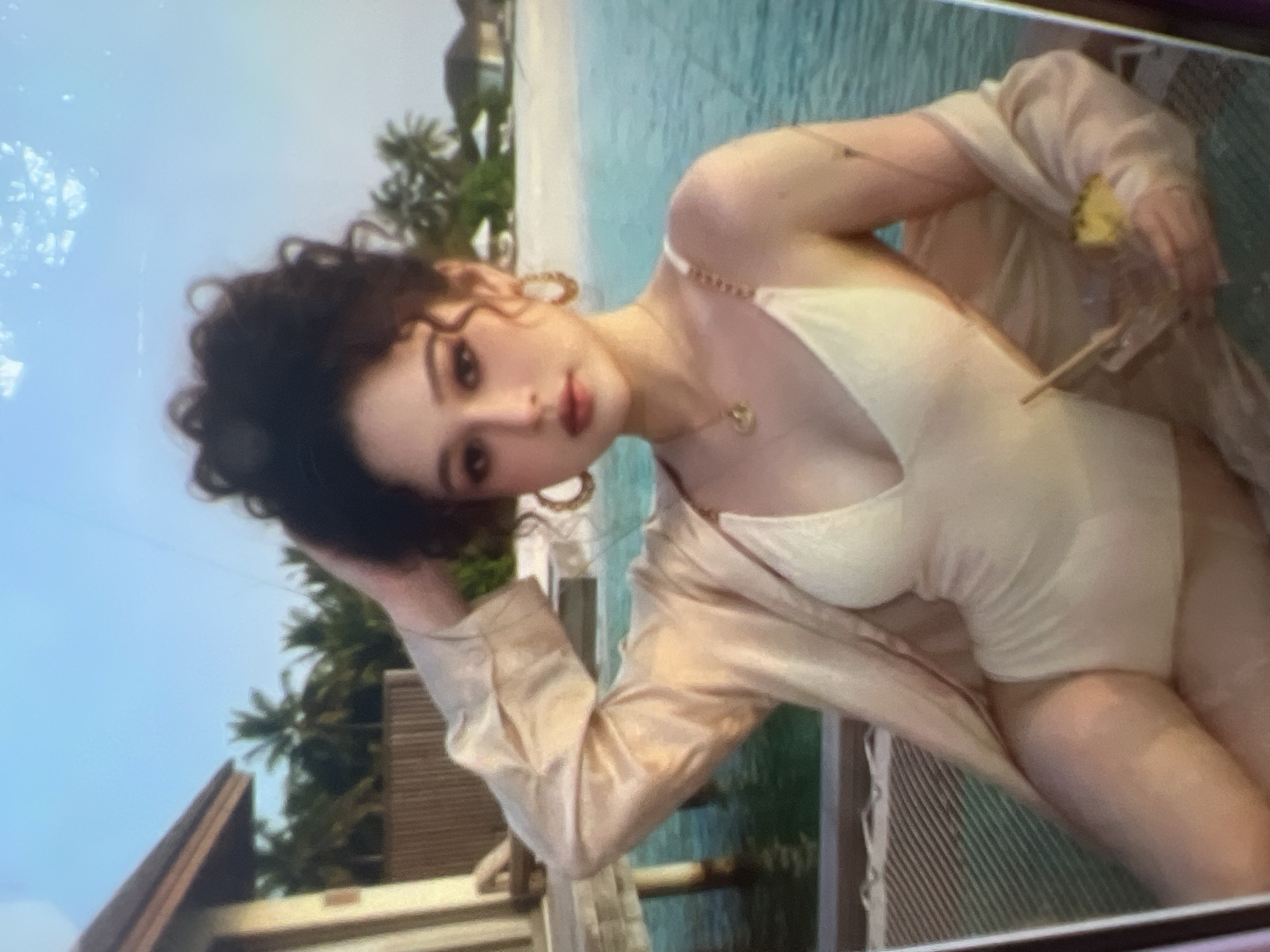 The image shows a young woman in a cream-colored swimsuit and a light-colored open robe, seated on a textured surface next to a body of water. She has dark, curly hair pulled up and is wearing large hoop earrings and a delicate necklace. In her right hand, she holds a drink with a straw and a slice of pineapple. Behind her, palm trees and small buildings are visible, suggesting a tropical or subtropical location. The water appears to be clear and blue. The scene is lit by natural light, likely from the sun. There is no indication of Kara-Dıykan, Kyrgyzstan in the image.