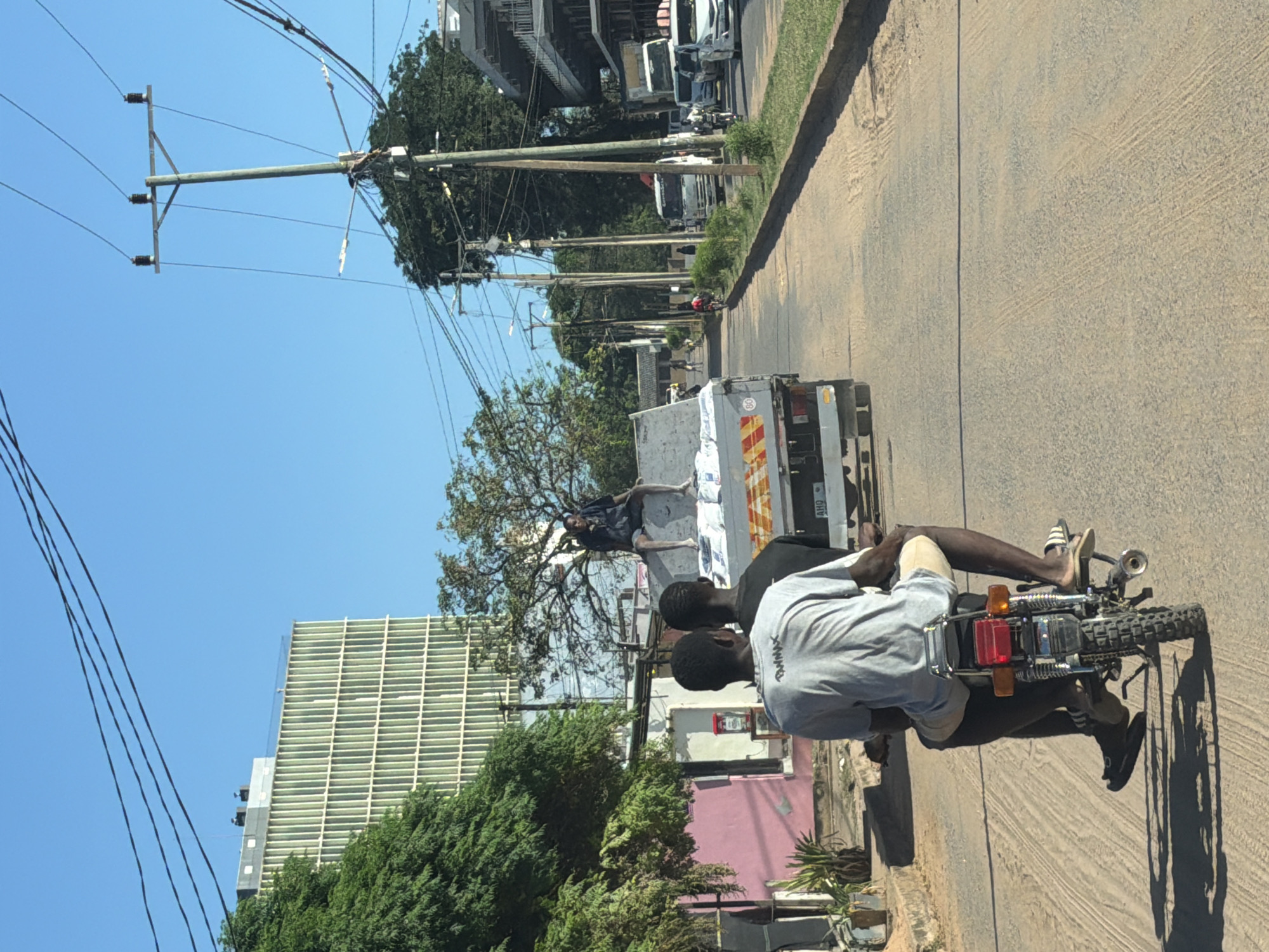The image depicts a bright, sunny daytime street scene in an urban or semi-urban environment. The perspective is from a vehicle, looking ahead.
In the immediate foreground, two men are riding a motorcycle on a paved road that appears somewhat dusty. The driver is wearing a light-colored short-sleeved shirt and light shorts, while the passenger, seated behind him, wears a darker shirt with the text "QWNX" visible on the back. Both individuals are wearing sandals or flip-flops.
Ahead of the motorcycle, a white pickup truck is traveling in the same direction. A man is casually seated on the open flatbed of the truck, facing away from the viewer. The rear of the truck is adorned with yellow and red striped reflective panels, a speed limit sign indicating "80" within a red circle, and a partial license plate or identifier that reads "AHO".
The right side of the road is bordered by a series of wooden utility poles, heavily laden with numerous overhead power lines that extend into the distance. Lush green trees and bushes are visible along the street, providing shade and greenery. In the midground and background, other vehicles, including trucks and cars, can be seen either moving or parked along the street. To the far left, a tall building with a distinctive modern, grid-like facade rises into the clear blue sky. The road itself shows signs of wear, and a dirt shoulder runs alongside it to the right.