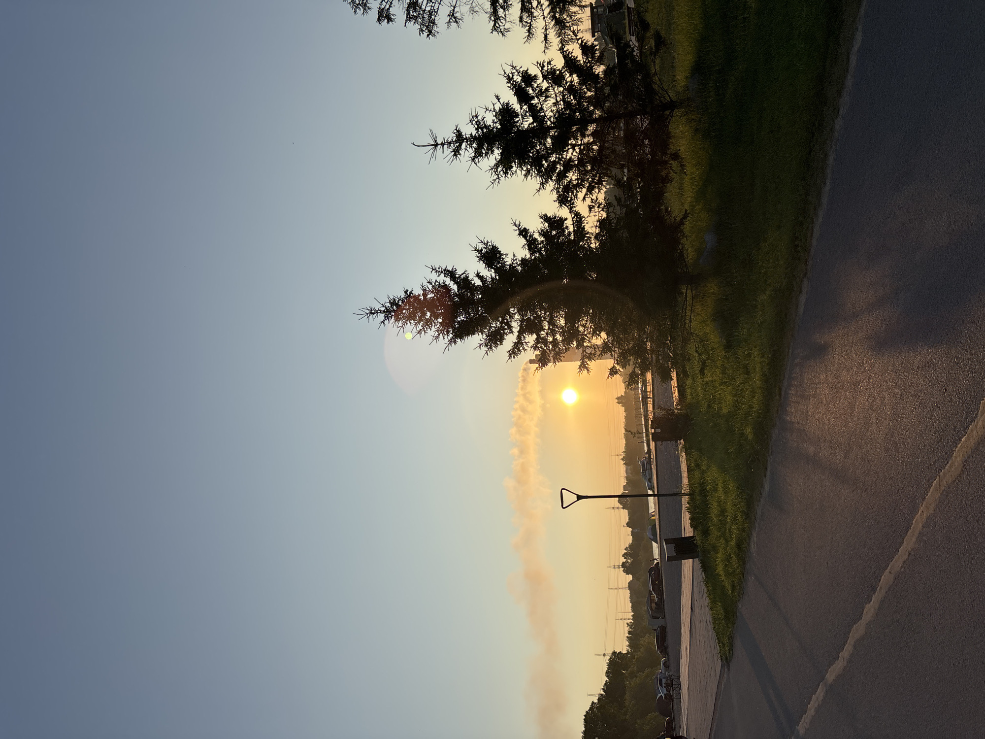 The image depicts an outdoor scene at sunset, likely in the city of Inskoy, Russia, given the context. In the foreground, a paved path runs diagonally from the bottom left to the middle right, with a yellow line marking its edge. A shadow of an unseen object falls across the path from the right. To the right of the path is a grassy area with several evergreen trees, silhouetted against the sky and the setting sun. One of the trees is in the mid-ground, and two others are slightly further back.
In the mid-ground, beyond the grassy area, is a body of water, possibly a river or lake, with a bridge or walkway crossing it in the distance. There are several cars parked along a paved area next to the water. Tents are visible near the water's edge, suggesting a campsite or gathering. A single street lamp with a distinctive triangular head stands on the paved area between the grassy section and the parked cars.
The sky is clear and exhibits a gradient of colors from a pale blue at the top to a warm yellow-orange near the horizon, indicating sunset. The sun itself is a bright, glowing orb positioned slightly to the right of the center, casting a warm light. A plume of what appears to be smoke or steam is visible in the sky to the left of the sun, colored orange by the sunset. In the far distance, power lines and pylons are discernible against the horizon. The overall impression is a serene, natural landscape at dusk, with signs of human activity like the path, cars, and tents.