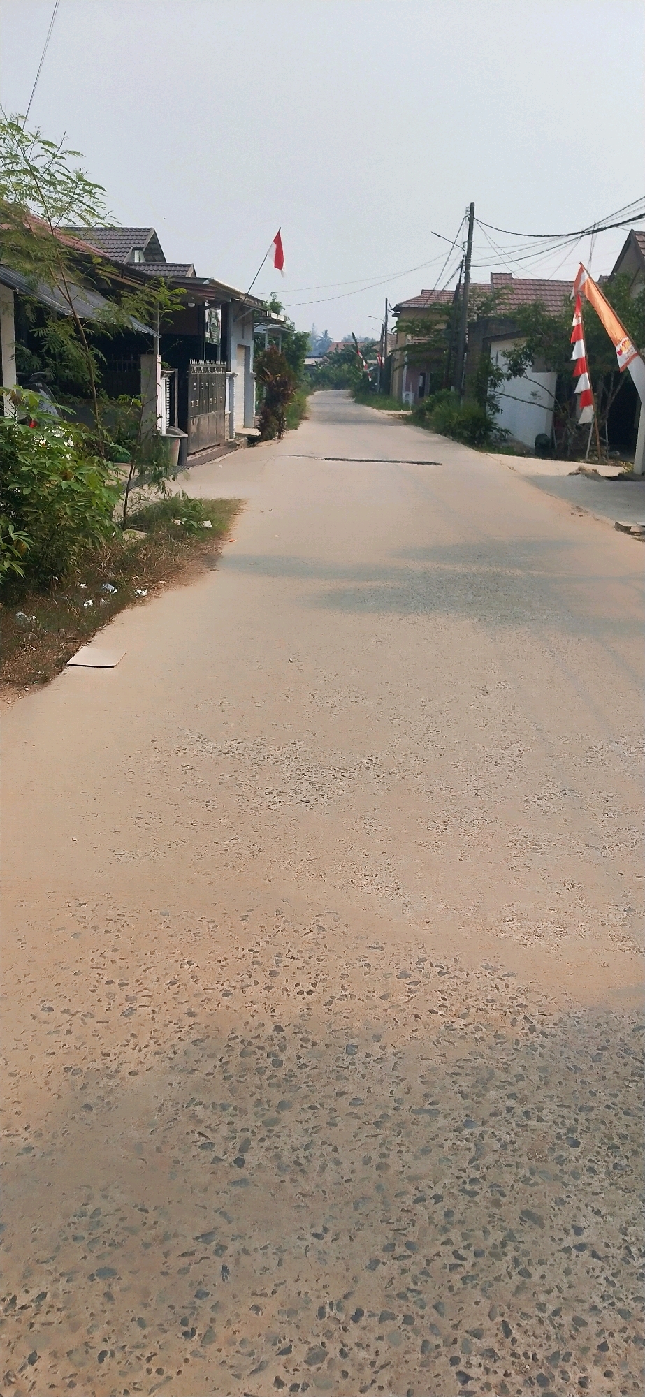 The image depicts a street scene in the city of Belimbing Raya, Indonesia. The road is paved with asphalt and appears to be in a state of disrepair with some cracked and worn sections. On either side of the road, residential houses with tiled roofs are visible. There are trees and vegetation bordering the properties. In the distance, the road curves and continues into what appears to be a residential area with more houses. Indonesian flags are displayed on poles along the street, one a simple red and white flag on the left and a more elaborate red and white striped flag on the right. The sky is overcast, suggesting a hazy or cloudy day. There are no people visible in the image. The scene is quiet and appears to be a typical day in a residential neighborhood in Indonesia.