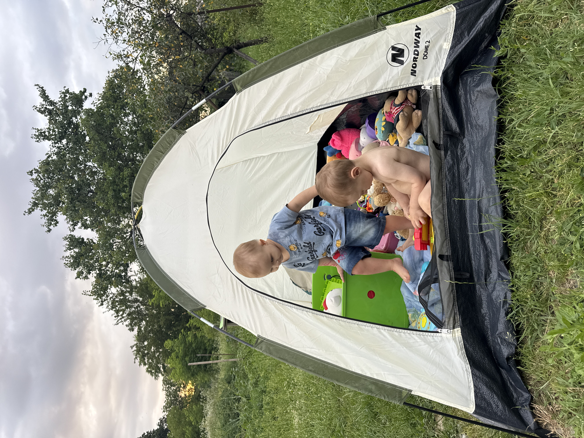 Two toddlers are inside a white and green Nordway Dome 2 tent pitched on a grassy area. The toddler on the left is wearing a blue t-shirt with Garfield on it and denim shorts, and is standing, reaching into a green plastic bin. The toddler on the right is shirtless and appears to be sitting and playing with toys. The tent is filled with stuffed animals and other toys. The background shows trees and vegetation, suggesting a rural or park-like setting. The sky is overcast with some hints of sunlight.
