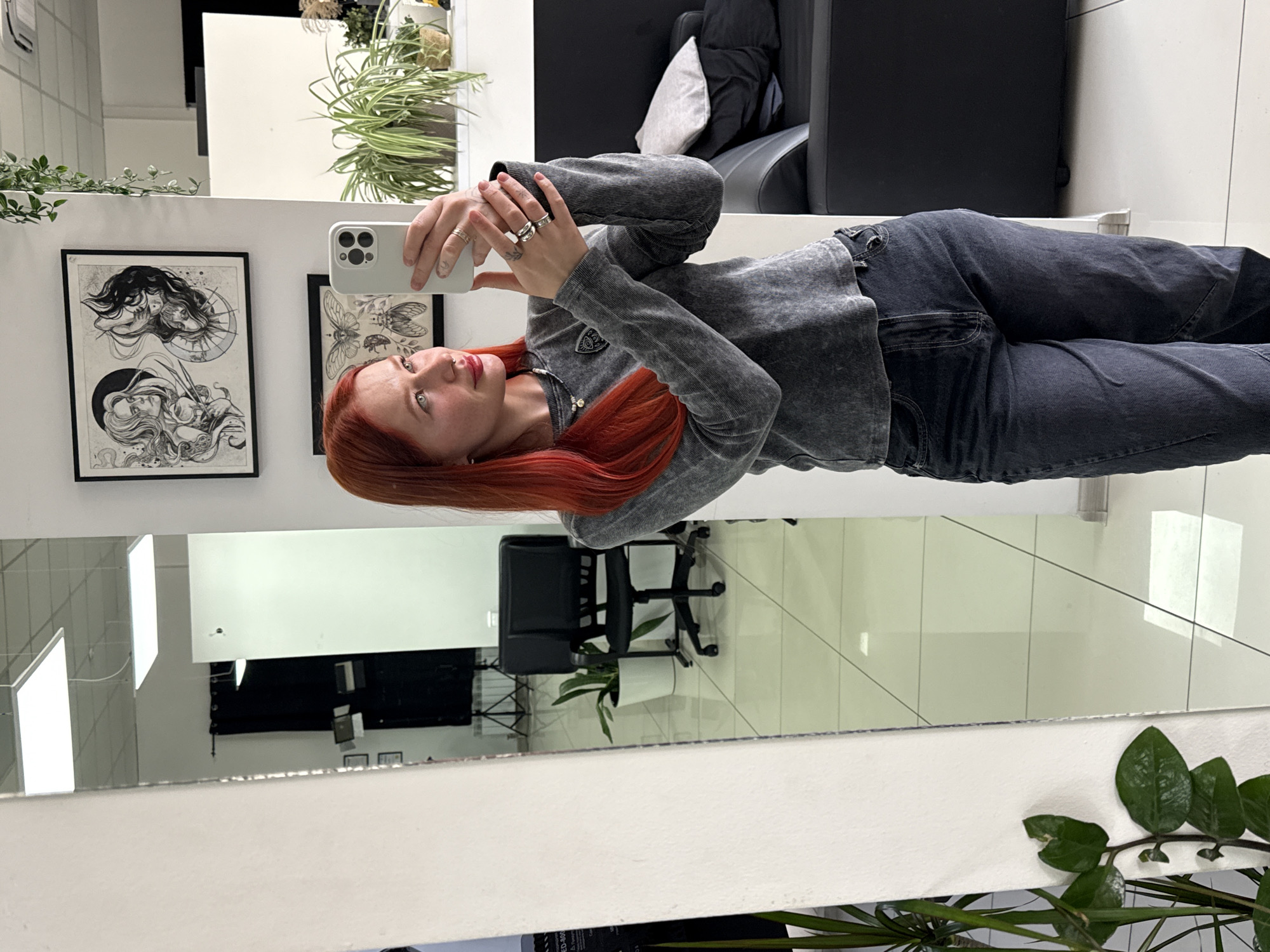 A woman with red hair is standing in front of a mirror and taking a picture with her phone. She is wearing a gray long-sleeved shirt and dark jeans. Behind her, there are framed art prints on the wall, plants, and a black chair. The reflection in the mirror shows an office space with chairs and plants. The location is not specified to be in the city of Rostov-on-Don, Russia, in the context of the image.