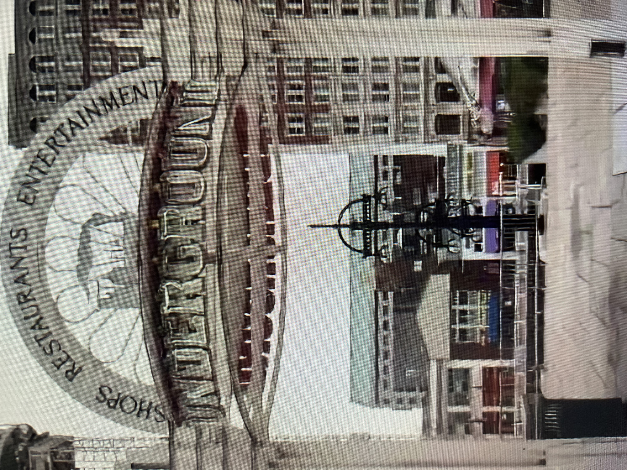 The media file shows a view of a modern cityscape with a focus on a large, circular sign. The sign is white and arched, with the words "SHOPS RESTAURANTS ENTERTAINMENT" inscribed in dark, serif lettering. In the center of the sign, there appears to be a stylized graphic or logo. To the right of the sign, vertical text is visible, though largely obscured and partially legible as "FOUNDERS..." The background consists of tall buildings with numerous windows, suggesting an urban environment. The architecture of the buildings appears to be a mix of older and newer styles. In the middle ground, there is a dark, intricate metal structure, possibly a sculpture or part of an architectural feature, with geometric elements. The lighting suggests daytime, but the sky is overcast, leading to a muted and somewhat desaturated color palette. No people are clearly visible in the frame. The overall impression is a still image of an outdoor urban scene, likely captured in a public space or plaza.