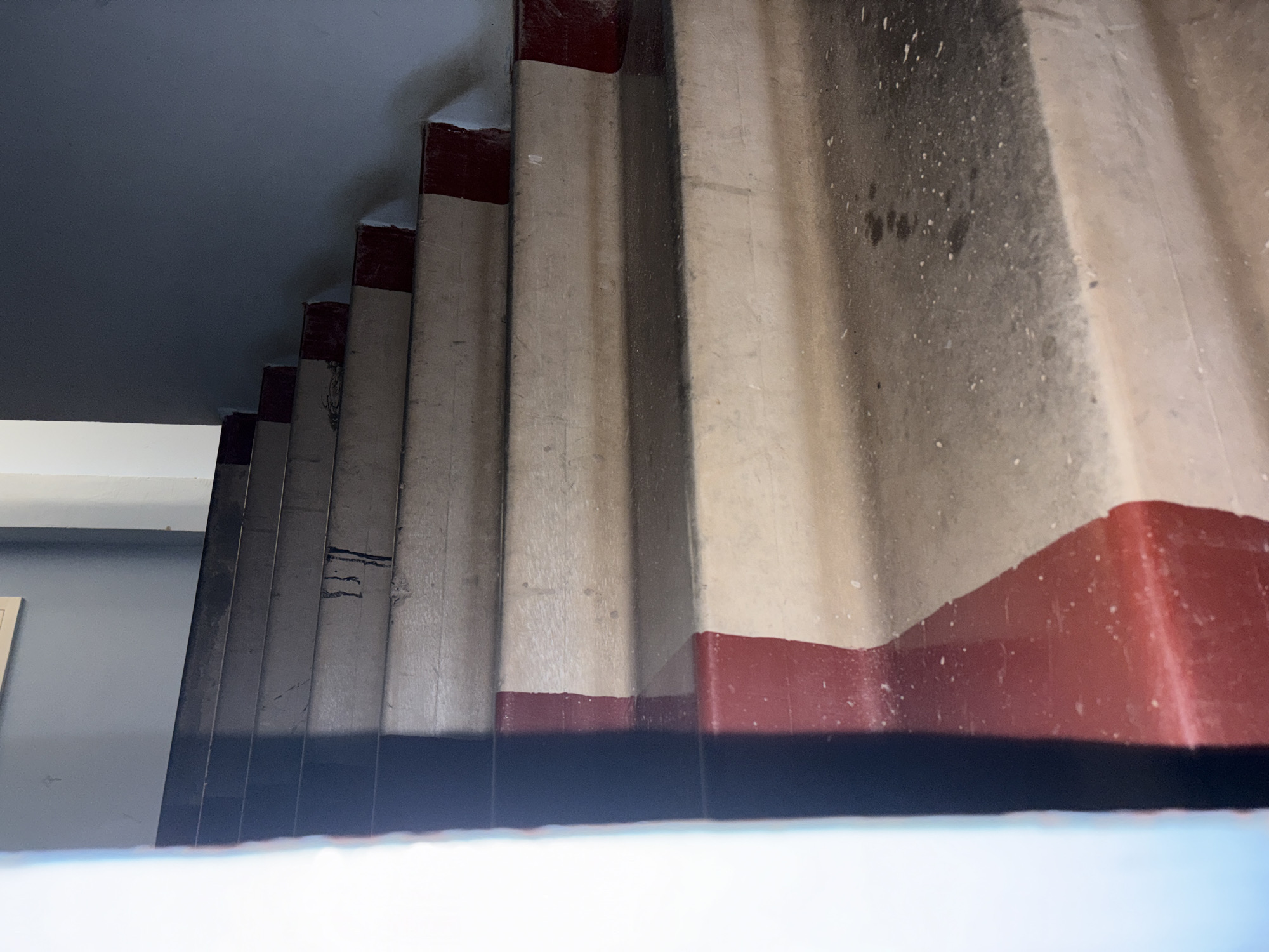 The image depicts an interior concrete staircase in Chaikovsky, Russia, viewed from a low angle as it ascends from the lower right to the upper left. The steps feature light beige-grey treads, with the risers and the outer edges of the treads painted a faded, dark red-maroon. The paint is visibly worn, chipped, and inconsistent in coverage across multiple steps. The concrete surfaces of the treads exhibit accumulated dirt, scuff marks, and areas of discoloration. Dark, vertically oriented streaks, consistent with liquid residue or heavy dirt, are present on one of the upper treads. The walls flanking the staircase are painted a dark grey-blue, with a brighter, lighter-toned vertical section visible along the far left edge of the frame. No individuals or active subjects are present in the scene.