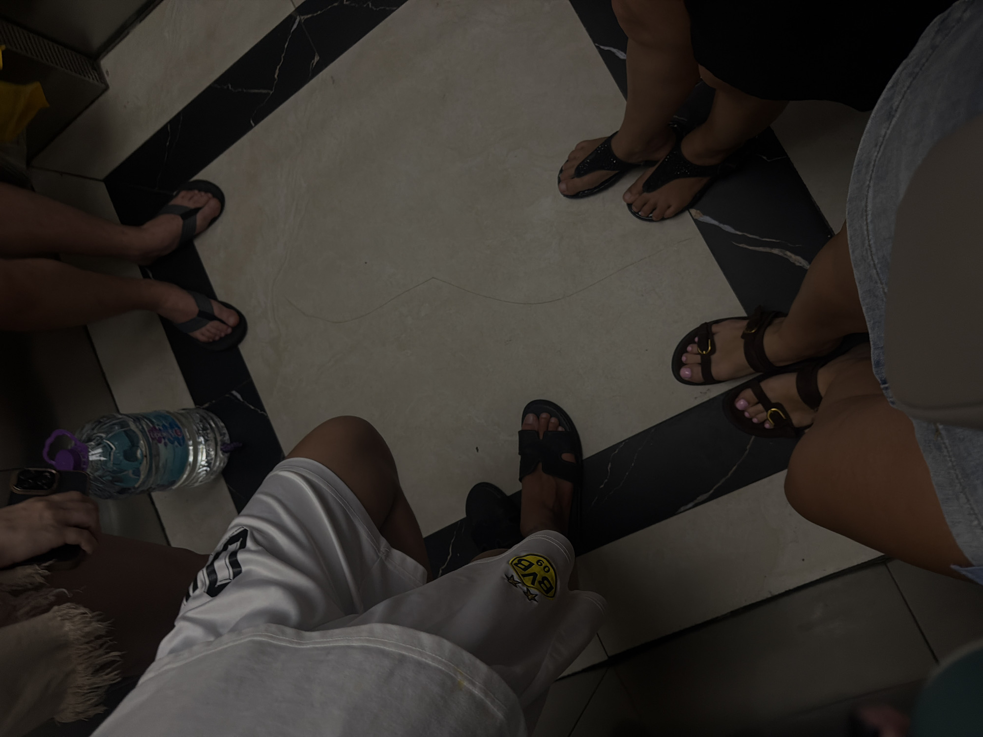 An overhead shot captures the lower legs and feet of at least five individuals gathered indoors, likely in Bac Nha Trang Ward, Vietnam. The subjects are arranged in a loose circle or semi-circle, their feet resting on a geometrically patterned floor composed of light, off-white tiles and darker, possibly black marble-effect tiles.
Various types of sandals and flip-flops are worn by the group. One person wears simple dark flip-flops. Another has black, possibly sparkly, thong sandals. A third pair of feet sports dark sandals with a prominent gold buckle, with light pink nail polish visible on the toenails. One person wears white shorts, which feature a distinct yellow circular patch with the text "BVB 09" and stars, indicating the logo of the football club Borussia Dortmund. This person also wears dark, possibly leather, slip-on sandals. Another individual wears denim shorts.
A plastic water bottle with a purple cap and a partially visible label, which appears to say "AQUA," is resting on the floor near one person's leg. A black smartphone is held in one hand, near the water bottle. The lighting in the scene is somewhat dim, suggesting an indoor environment or possibly evening, contributing to a relaxed and casual atmosphere. The activity appears to be a social gathering, possibly a break or waiting period, with people seated or standing in proximity to each other. No specific weather conditions are discernible, and the exact time of day is not clear from the image.