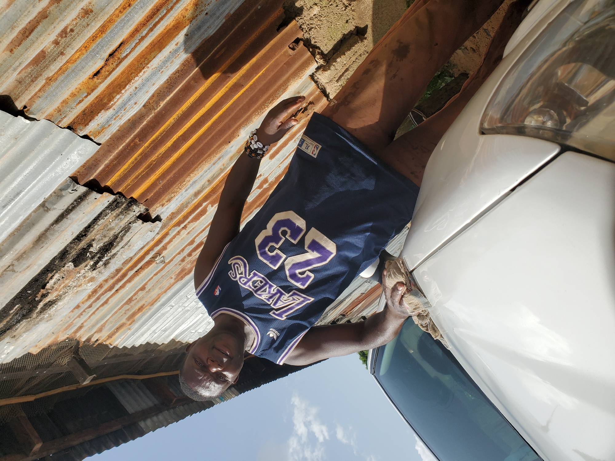 In Bukuru, Nigeria, a man wearing a blue Los Angeles Lakers jersey with the number 23 and brown pants is seen cleaning the hood of a white car with a tan cloth. He is standing next to the car, which is partially visible on the left side of the frame. Behind him, there is a structure made of corrugated metal roofing that appears weathered and rusty. The ground is dirt, and some dry vegetation is present. The sky is visible in the background with scattered clouds. The overall scene suggests an outdoor setting in a somewhat informal or developing urban environment.