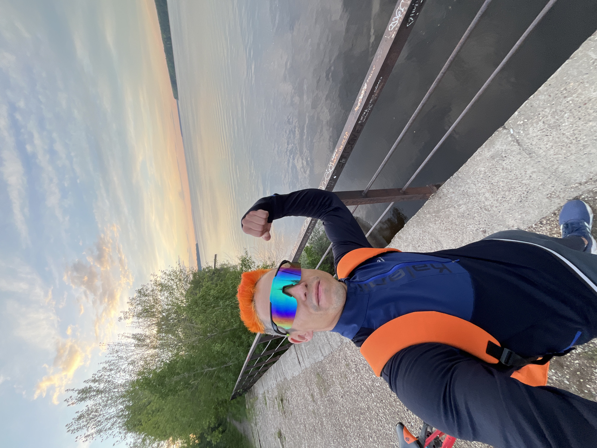 The image shows a man with orange hair and colorful sunglasses standing on a concrete embankment next to a body of water. He is wearing a dark long-sleeved shirt, dark pants, and blue running shoes, with an orange safety belt or life vest around his waist. His right arm is raised and flexed. A metal railing separates the embankment from the water. The sky is visible in the background, displaying a sunset or sunrise. Green trees grow along the bank further in the background. Graffiti is present on the metal railings.
Given the city is Chaikovsky, Russia, and the presence of a body of water, it's likely a location along the Kama River or one of its tributaries. The exact location requires further information.