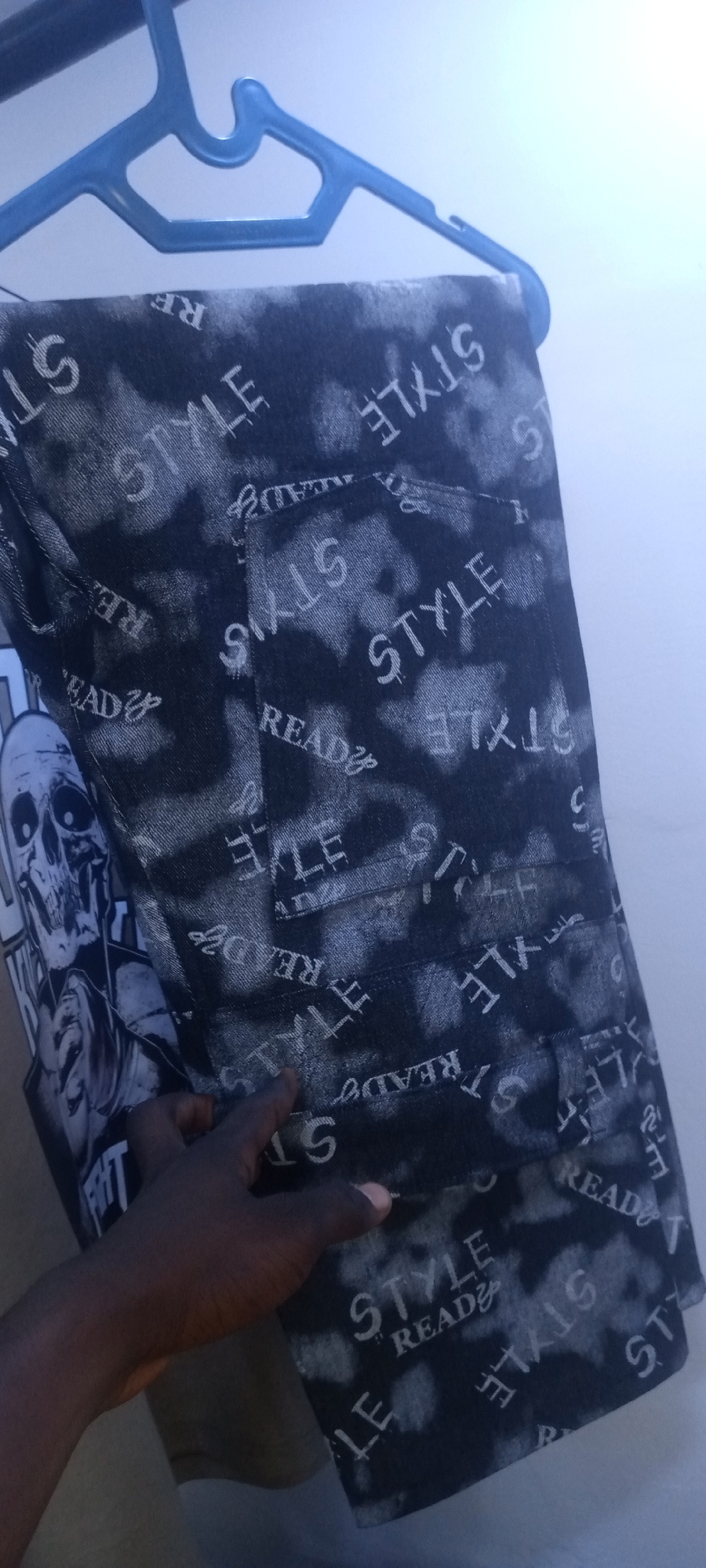 A pair of black and white patterned jeans hangs from a blue plastic hanger against a white wall. The jeans are patterned with the words "STYLE" and "READY." A dark-skinned hand touches the jeans near a T-shirt with a graphic print of a skull. The photo's location is unknown, and there's no specific evidence to indicate Jos, Nigeria.