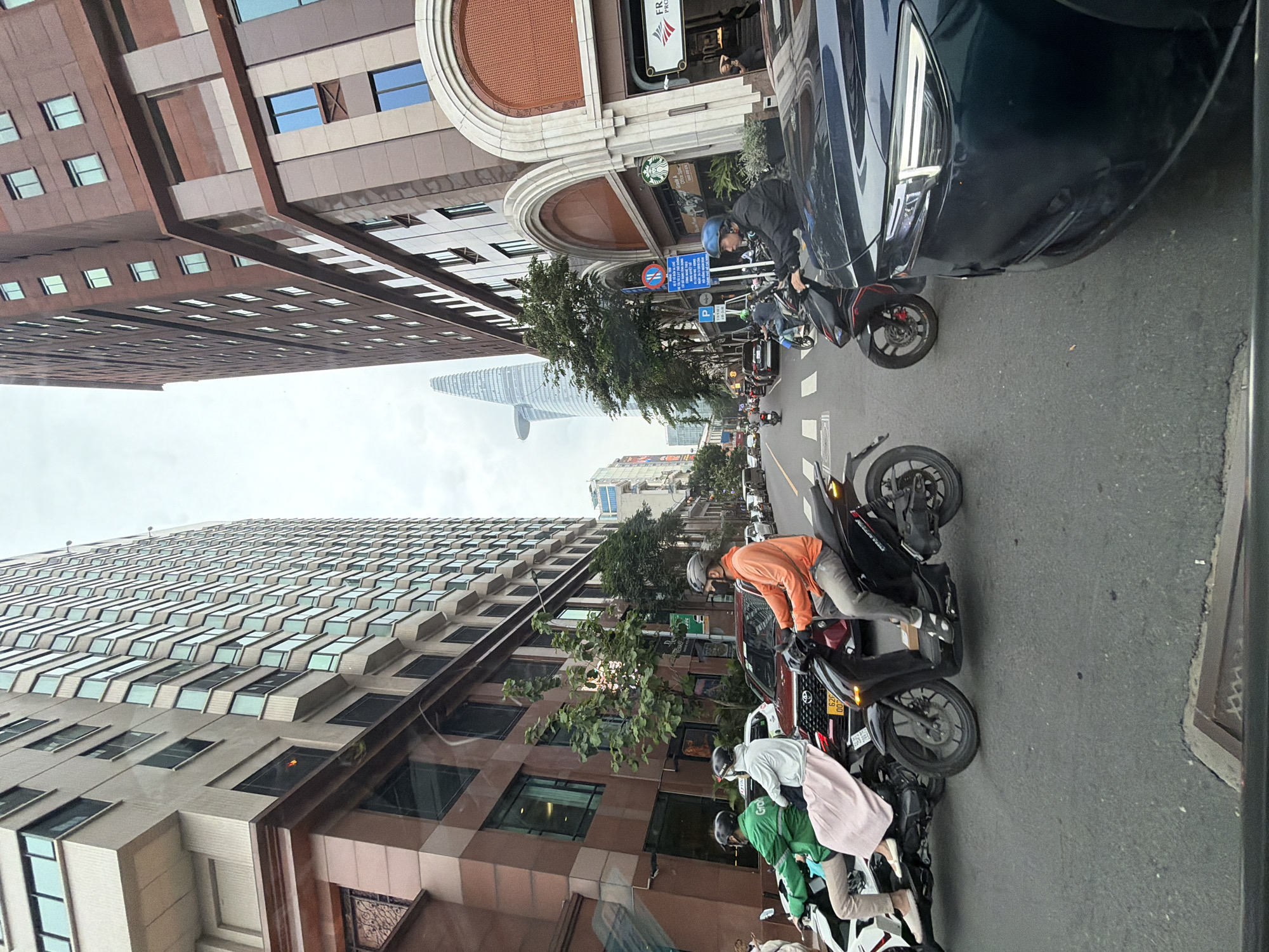 The image captures a street scene in Thủ Đức, Vietnam, characterized by tall, modern buildings and moderate traffic, primarily consisting of motorcycles. On the left, a light-colored building with numerous windows dominates the frame. To the right, a taller, darker brick building with arched entrances and a visible Starbucks logo stands prominently. In the foreground, several motorcycles are present. One motorcycle, ridden by a person in an orange shirt, is in the center, while another, ridden by a person in a green jacket and a passenger in a pink skirt, is on the left. A dark-colored car is parked or moving in the foreground on the right. The sky is overcast, suggesting a cloudy day. The presence of multiple motorcycles and the architectural style suggest an urban environment in Southeast Asia. The distinctive skyscraper in the background, with its unique twisting shape, is likely Landmark 81, a prominent building in Ho Chi Minh City, which Thủ Đức is part of.