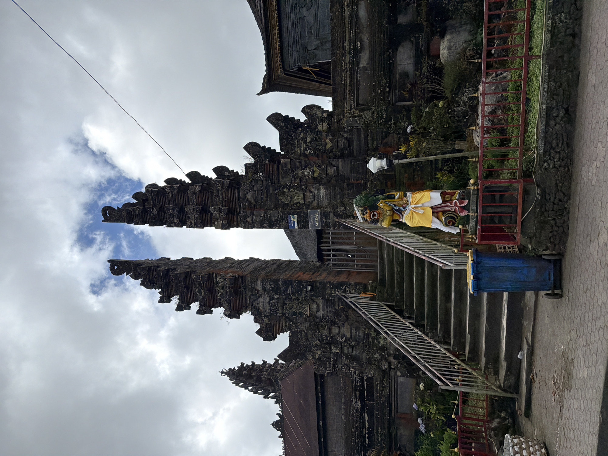 This media file captures a portion of a traditional Balinese temple complex in Kintamani, Indonesia. The scene is dominated by weathered stone architecture, characterized by multi-tiered pagoda-like structures with ornate carvings and mossy surfaces. In the foreground, a stone staircase with metal railings leads upwards, suggesting an entrance or pathway within the temple grounds.
At the base of the stairs, a vibrant statue or effigy stands, adorned with yellow and white fabric. This figure is likely part of a religious or cultural representation specific to the temple. To the right of the stairs, a blue trash bin on wheels is visible, hinting at the ongoing use and maintenance of the site. The surrounding area features lush greenery and other stone elements, typical of Indonesian temple architecture.
The sky above is a mix of white and grey clouds, with patches of blue visible, indicating an overcast or partly cloudy day. The lighting suggests it is daytime, though the exact time is not discernible. The overall mood of the image is one of historical significance and cultural immersion, with the ancient stone structures conveying a sense of timelessness.
There are no clear indications of people actively engaged in religious ceremonies or specific events, but the presence of the statue and the infrastructure suggests an active place of worship or tourism. The image is vertically oriented. There is some text visible on a sign behind the statue, which appears to read "MEDAL".