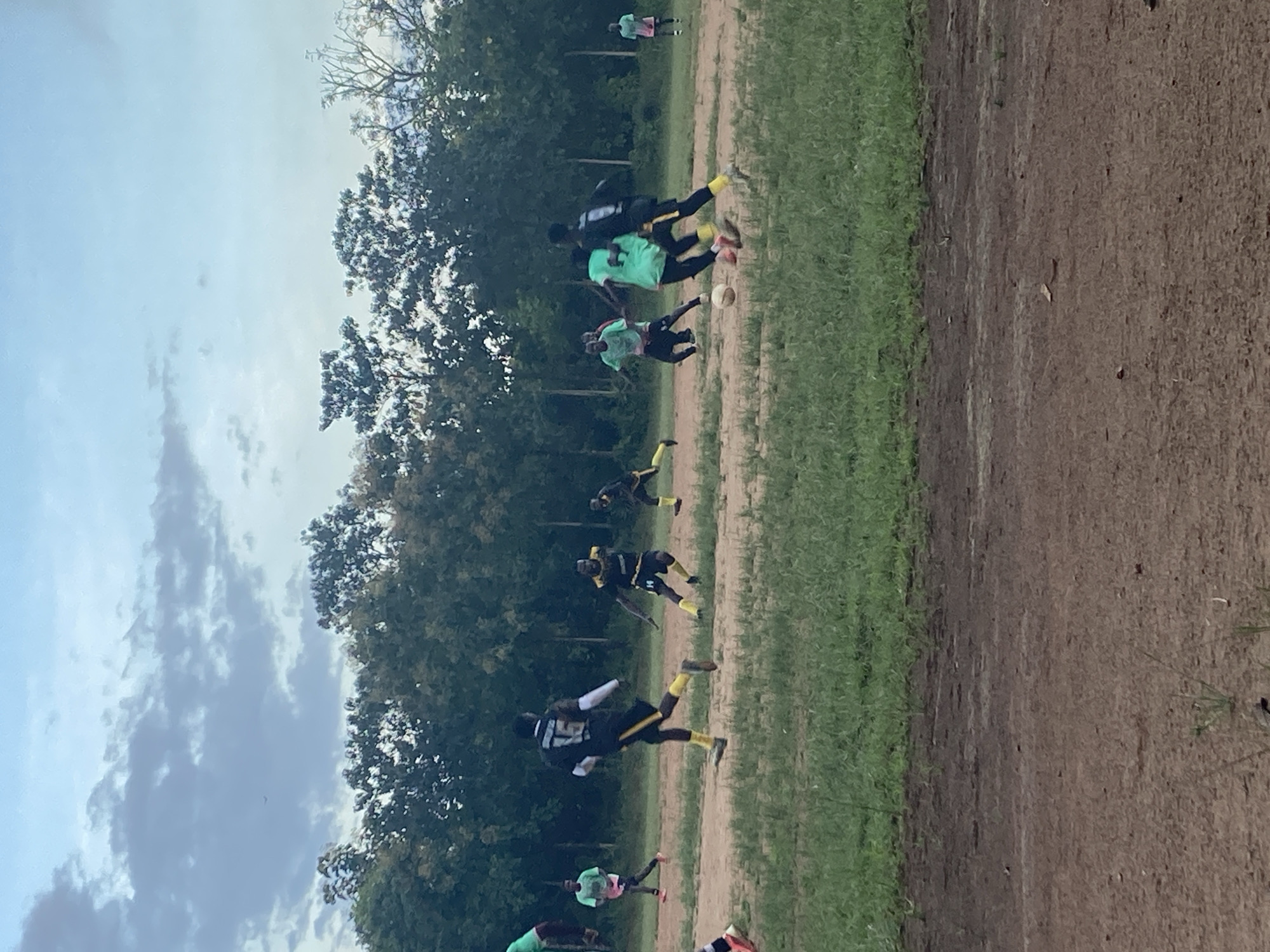 The image depicts a group of young men playing soccer on a dusty, uneven field surrounded by sparse green grass and a line of trees in the background. The sky above is cloudy with hints of blue. Several players are in motion, actively engaged in the game. One player in the foreground wears a black jersey with the number "15" and yellow trim. Another player in a green jersey is closely contesting for the ball. The general setting suggests an outdoor recreational activity in a semi-rural or less developed urban area. The presence of a soccer game implies community or leisure activity within Miango, Nigeria.