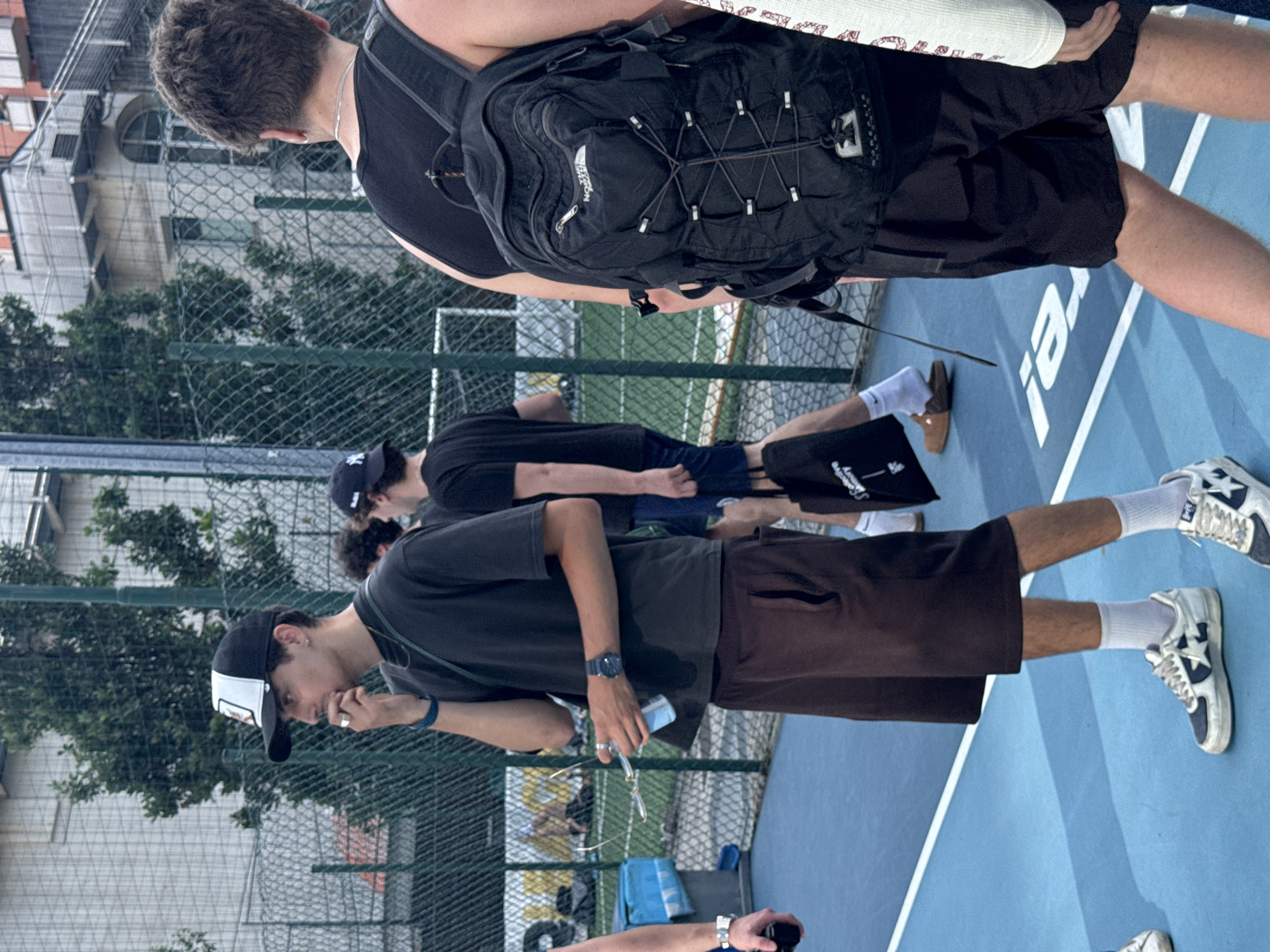 The image depicts an outdoor scene on a blue sports court in Milan, Italy, bordered by a green chain-link fence. Several young men are present. The central figure, a young man with dark hair and a mustache, wears a black and white baseball cap, a dark grey short-sleeved t-shirt, and dark brown knee-length shorts. He has a thin gold chain, a blue bracelet on his left wrist, and a dark watch on his right. His left hand is raised towards his mouth, holding a small object, while his right hand holds a pair of sunglasses and a small clear bottle. He wears white socks and light-colored sneakers with a dark star design.
To the right, a young man seen from the back has short curly brown hair and wears a black tank top, black shorts, and a black North Face backpack with bungee cords. He wears a thin silver chain and a silver hoop earring. Partially visible on his feet are white socks and sneakers with a star design similar to the central figure's.
Behind the central figure, another young man is partially visible, wearing a dark baseball cap with a "NY" logo and a dark t-shirt, carrying a black tote bag. On the far left, an arm with a silver watch holds a black object. The blue court surface features white lines and partially visible white lettering that reads "re!" (reversed). In the background, beyond the fence, are trees and multi-story buildings, one with a light-colored facade and external stairs, and another with a reddish-brown facade.