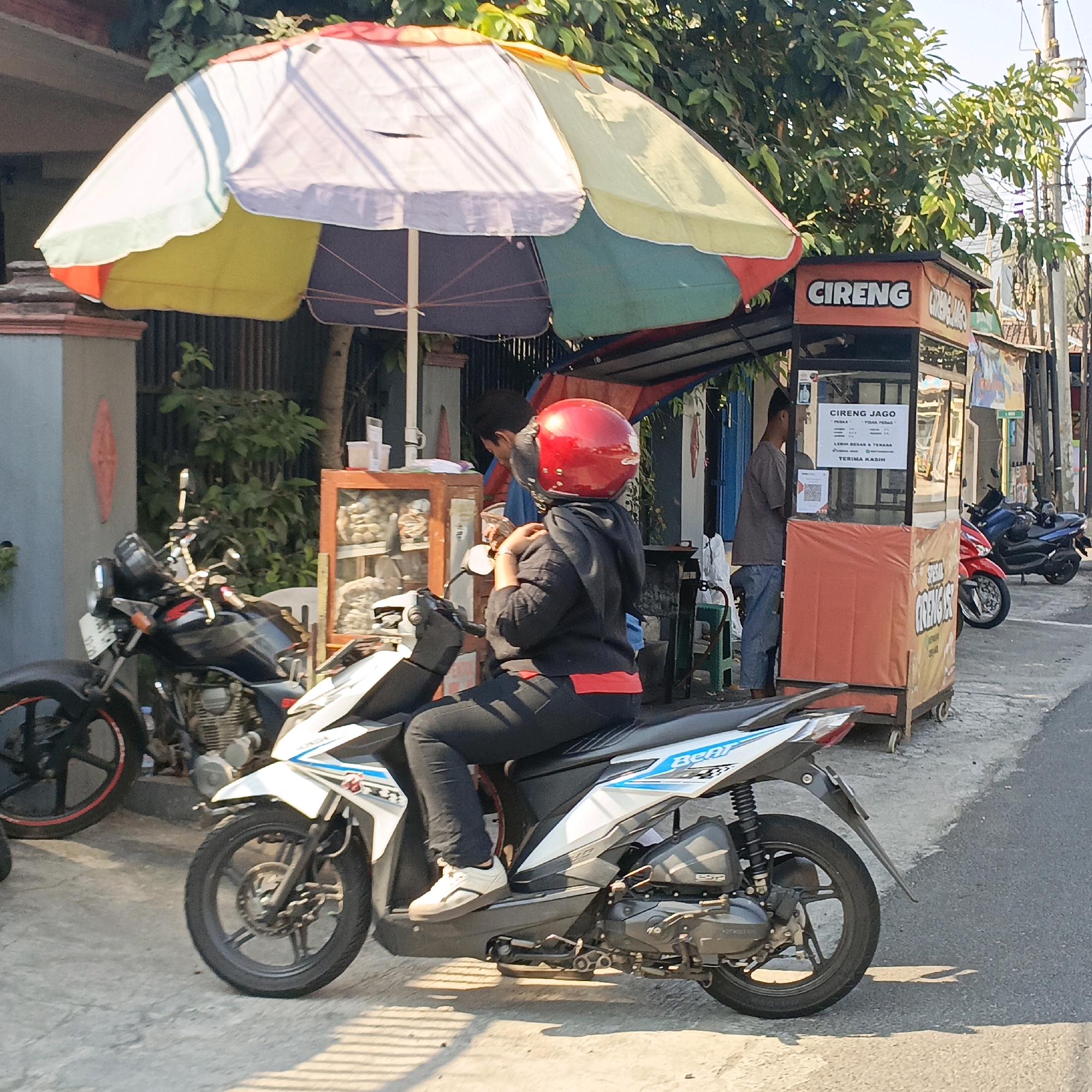 A street scene in Salatiga, Indonesia, features a person wearing a red helmet and a black hoodie, seated on a white and blue scooter. The scooter is parked on a concrete surface next to a food stall. The stall, adorned with an orange and yellow awning, displays a sign that reads "CIRENG JAGO" and lists prices for various items. In the background, other food stalls and buildings are visible, along with several parked motorcycles, including a black one to the left of the scooter. The environment is illuminated by sunlight, casting shadows on the ground.