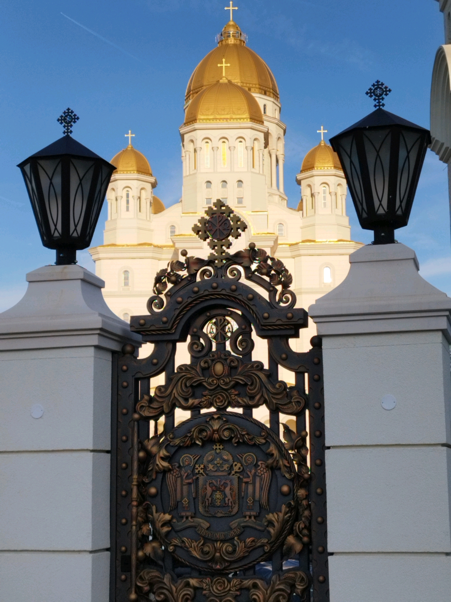The image captures an exterior view of the Cathedral of the Salvation of the Romanian People in Bucharest, Romania. In the foreground, a large, ornate, dark metal gate stands prominently. The gate features intricate scrollwork and a central emblem that appears to be a coat of arms. Flanking the gate are two white stone pillars, each topped with a black, hexagonal lantern. These lanterns have decorative metal cages and are crowned with a black Orthodox cross.
Behind the gate and pillars, the white façade of the cathedral rises, punctuated by rows of windows and topped with multiple golden domes, each also adorned with a cross. The sky above is a clear, bright blue with a faint white contrail from an aircraft visible in the upper left quadrant. The lighting suggests it is daytime, likely morning or late afternoon, given the clear sky and the direction of light on the cathedral. The scene is static and devoid of any people.