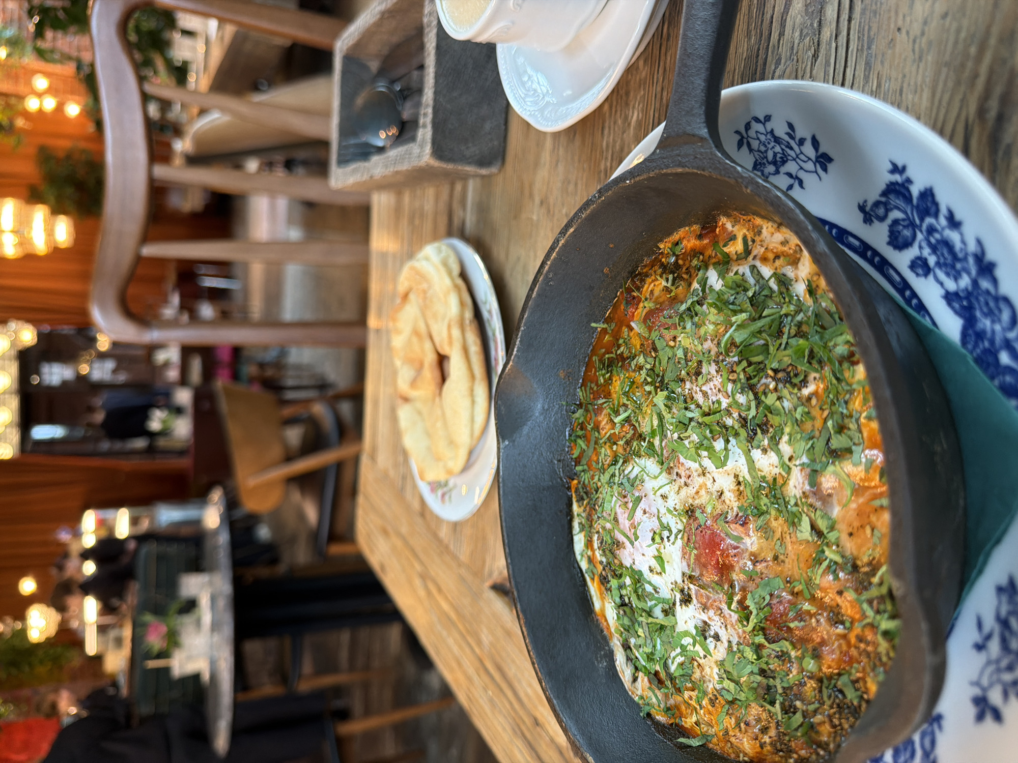 The image depicts a close-up view of a meal served in a restaurant, likely located in Saint Petersburg, Russia. In the foreground, a black cast-iron skillet contains a dish with eggs cooked in a tomato-based sauce, garnished with fresh green herbs. The skillet rests on a decorative white plate with blue floral patterns. To the right of the skillet, a white cup with a beverage, likely coffee or tea, sits on a saucer. A small wooden box containing cutlery, specifically a spoon, is also visible. In the background, a round table with chairs is set up in a dimly lit dining area, with patrons blurred in the distance and warm lighting from chandeliers and wall sconces. The overall ambiance suggests a cozy and intimate dining experience.