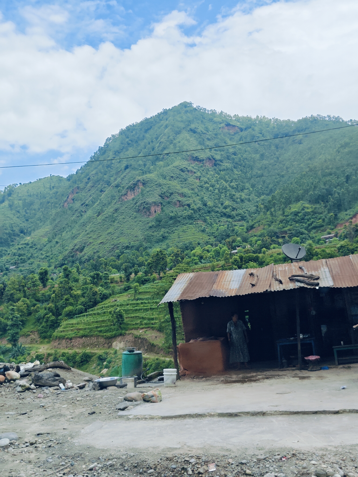 The image shows a mountainous landscape with a small, rustic dwelling in the foreground. The dwelling has a corrugated metal roof and appears to be made of mud and brick. A woman in a patterned dress is standing inside the dwelling. To the left of the dwelling, there are several large rocks, a green water tank, and some debris. In the background, the mountain is covered in lush green vegetation, with visible terraced fields. The sky is partly cloudy. The scene is likely located in a rural area such as Galchhi, Nepal, given the mountainous terrain and the style of the dwelling.