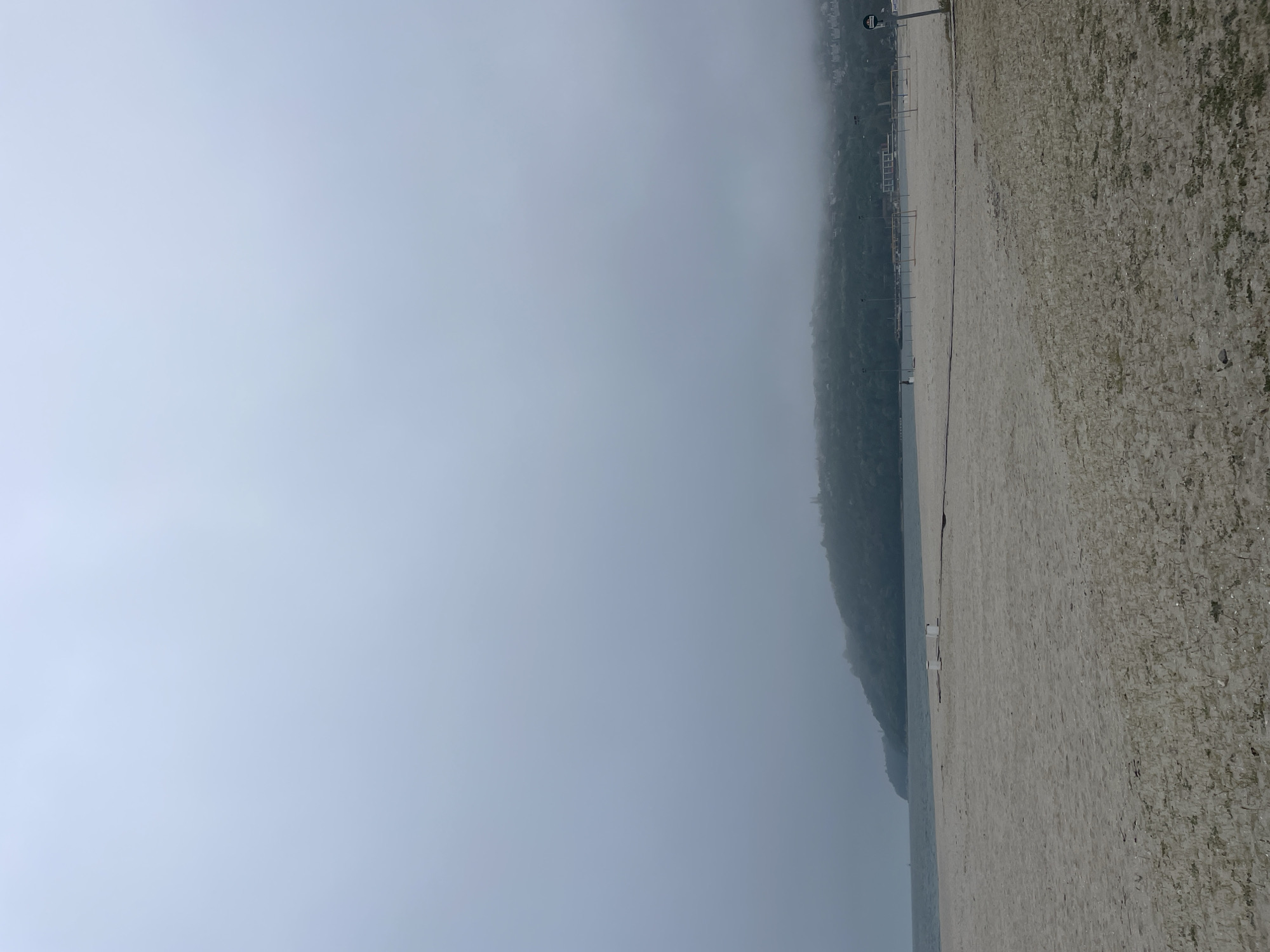 The image captures a beach scene in Varna, Bulgaria, shrouded in dense fog. The shoreline occupies the right side of the frame, covered with sand interspersed with small pebbles and vegetation. The sand appears damp, likely due to the fog. The sea is barely visible as a thin line due to the heavy fog. Along the beach, structural elements such as poles and possibly volleyball nets can be seen. A wooded hill or cliff looms behind the beach, mostly obscured by the fog, which also obscures any buildings or structures that might be on the hill. The predominant color is gray, due to the overcast conditions.