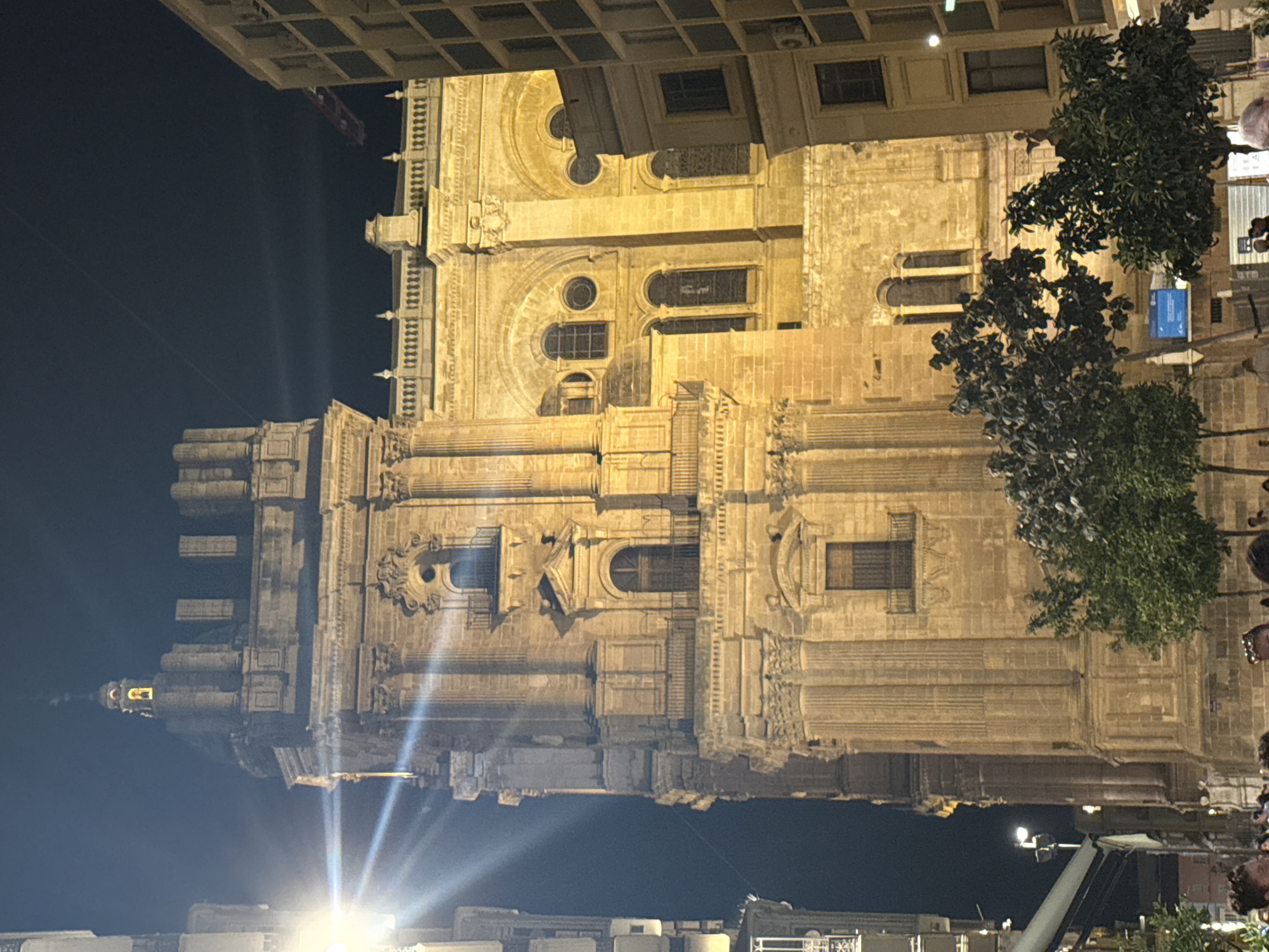 The image captures a nighttime view of the Málaga Cathedral in Spain. The imposing stone facade of the cathedral is illuminated by warm artificial lights, highlighting its intricate architectural details, including columns, arched windows, and decorative carvings. The building's grandeur is emphasized by the dramatic play of light and shadow.
In the foreground, trees with dense foliage partially obscure the lower sections of the cathedral. The sky above is dark, indicating that it is nighttime. The image appears to be taken from a street level, with some modern lighting fixtures visible at the bottom, including a bright light source casting strong rays.
The scene evokes a sense of awe and historical significance. The architecture suggests a significant religious or historical building, typical of European cities. The presence of the trees adds a natural element to the urban landscape. No people are clearly discernible in the image, and the overall focus is on the magnificent structure of the cathedral. The clear sky suggests no particular adverse weather conditions. A blue sign is partially visible on the right side of the image, but the text is not legible.