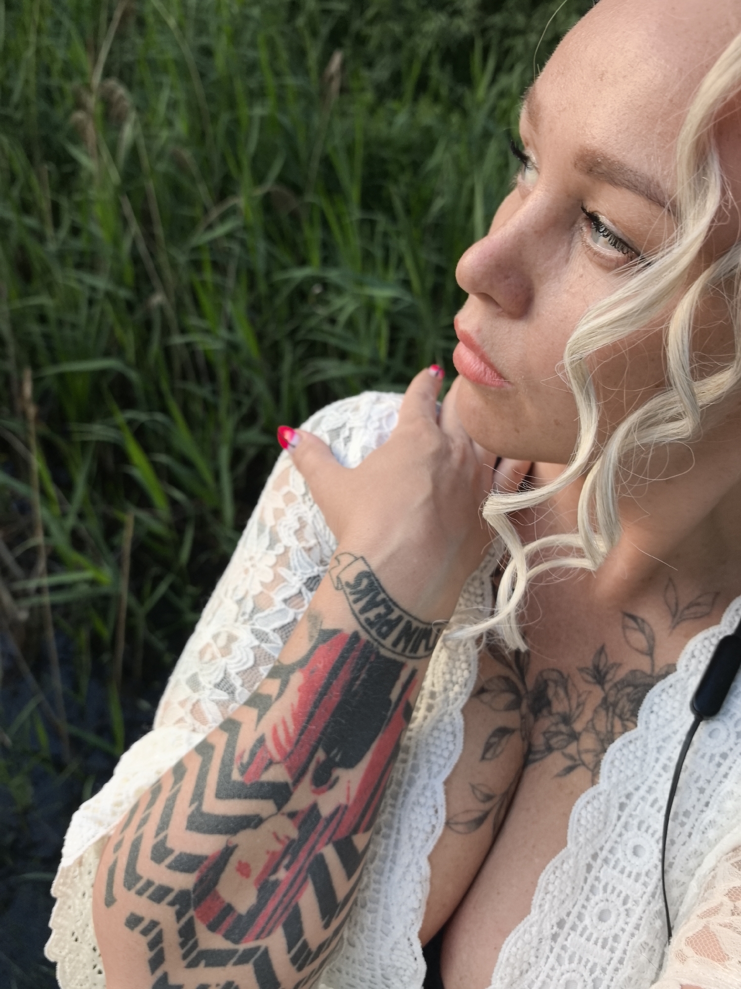 A medium close-up shot shows a fair-skinned woman with blonde curly hair, seen from the side, looking upwards and to the right. Her face is partially in shadow. She is wearing a white lace top. Her left arm is visible, covered in tattoos, including a black and red geometric design and text. Her right hand is near her face, with red nail polish. The background is out of focus and appears to be tall, green grass and water, suggesting an outdoor setting, possibly near a marsh or a lake in Noviy, Russia.