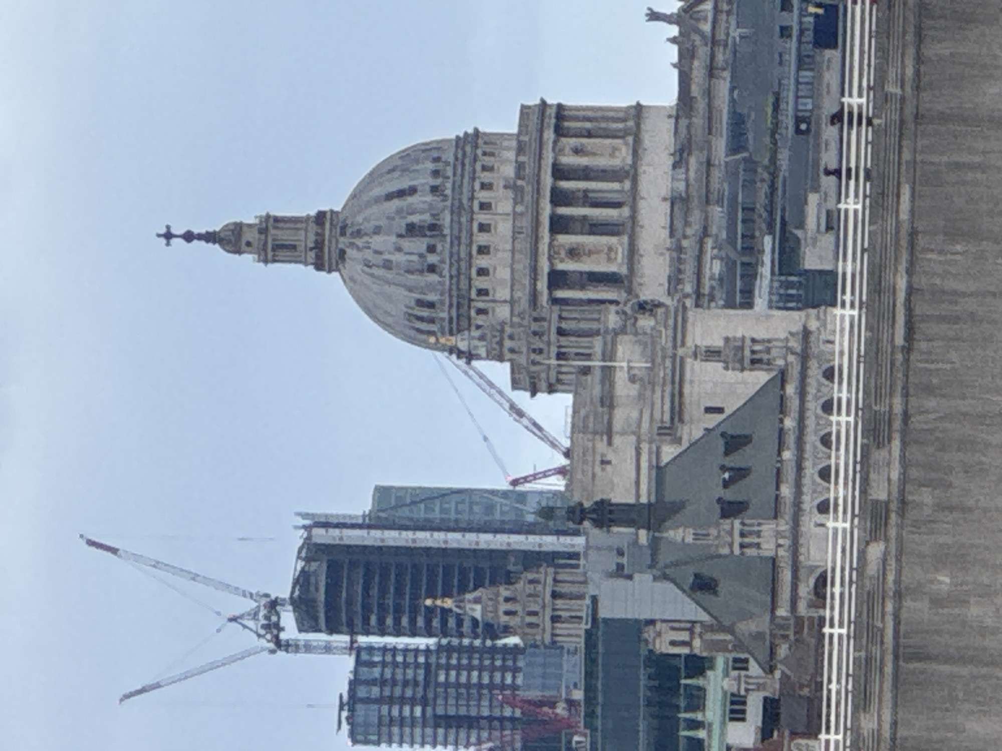 The image shows St. Paul's Cathedral in London, United Kingdom, seen from an elevated position, possibly the Tower Bridge walkway. The cathedral's dome is prominent, with details of its layered structure and cross at the top visible. Several modern high-rise buildings are present in the background, some under construction, indicated by cranes. The perspective is slightly skewed, making the scene appear somewhat tilted. The sky is overcast, casting a muted light over the urban landscape.