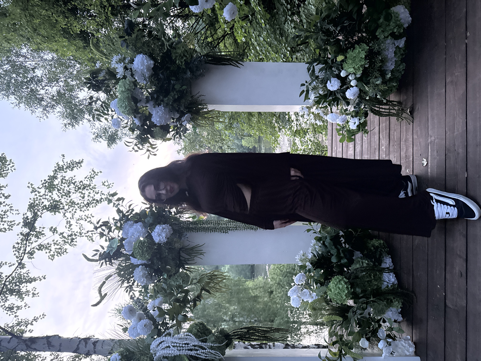 A person is standing on a wooden deck in what appears to be a park or garden setting. The person is wearing dark clothing and sneakers with white stripes. Decorative white flower arrangements are placed on and around white pedestals or archways. The background features green foliage and trees, possibly near a body of water. The location is indicated as Эстачи, Russia.