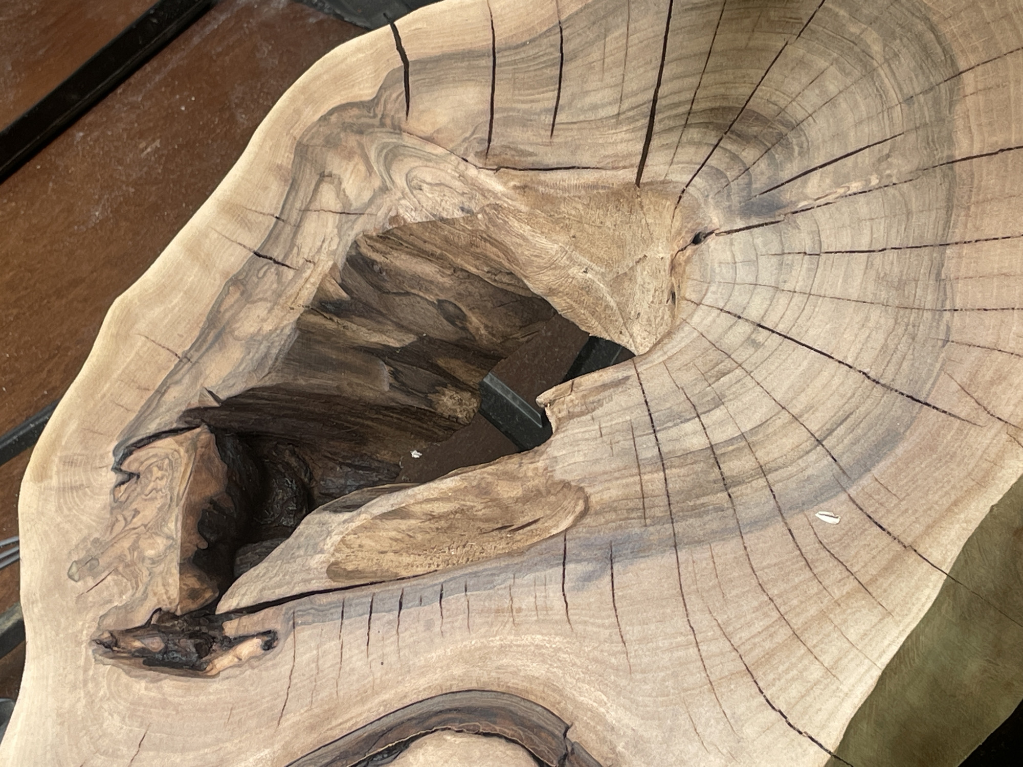 A close-up, overhead shot of a live-edge wooden tabletop with a large, irregular void in the center. The wood grain is prominent, with visible growth rings and dark, crackled lines. The void is deep and dark, with rough, uneven edges. A dark, rectangular object, possibly a tool or a piece of hardware, is partially visible within the void. The tabletop appears to be resting on a dark surface. The image does not provide any information about the city of Stryzhavka, Ukraine.
